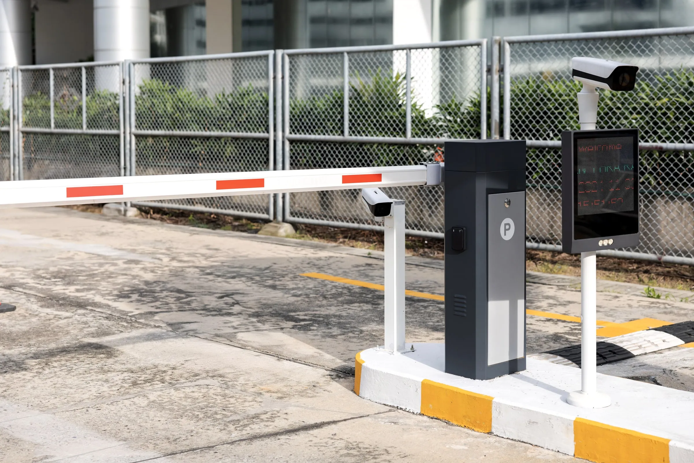 Automatic barrier arm at a parking entrance with a camera and a display post, chain-link fence and office building behind