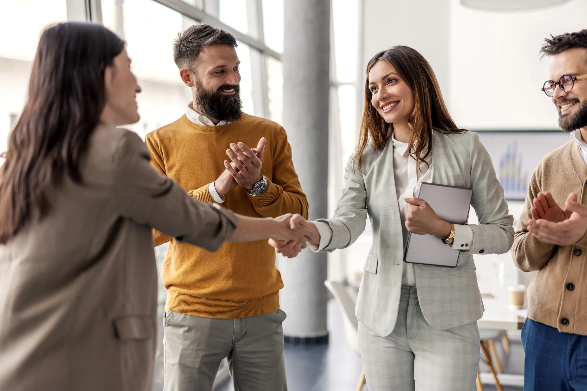 Businesswoman holding a tablet shaking hands with a colleague in a bright office while two coworkers applaud