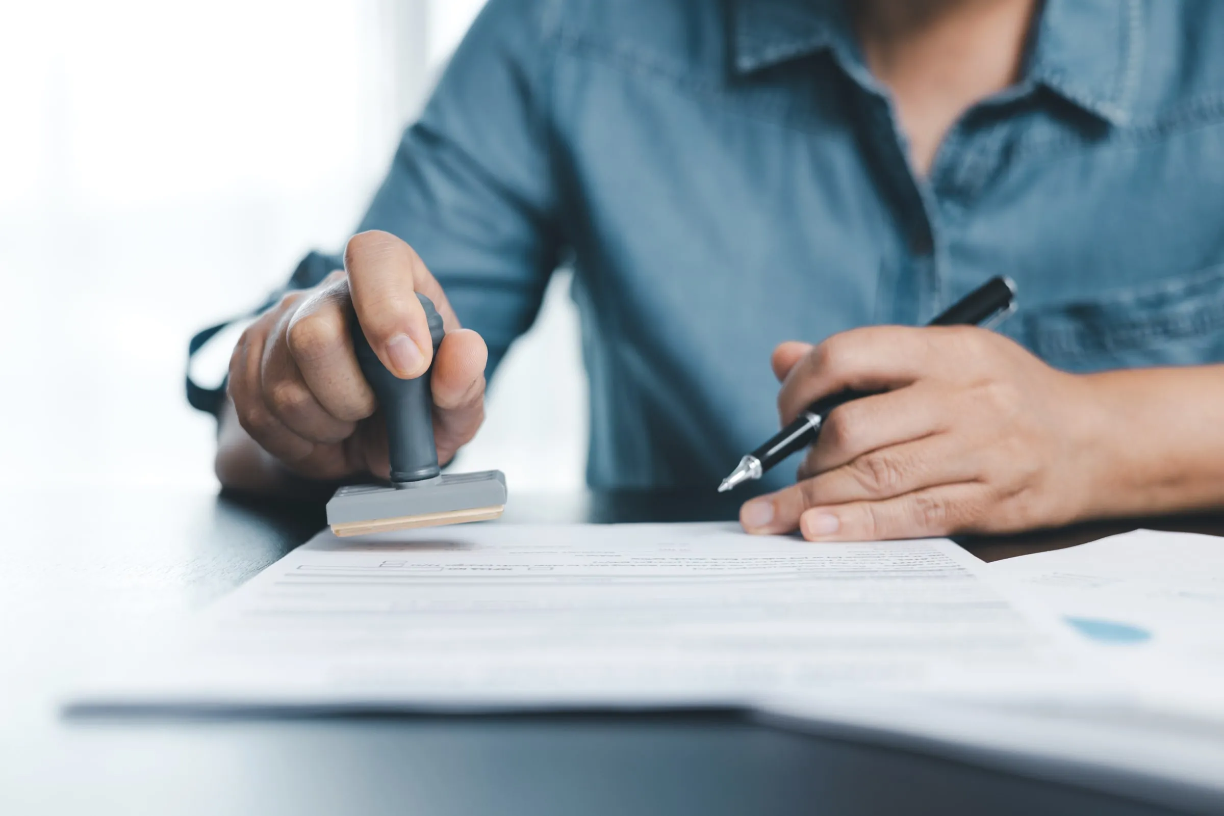 Close-up of a person in a blue shirt stamping an approval on a printed form while holding a pen