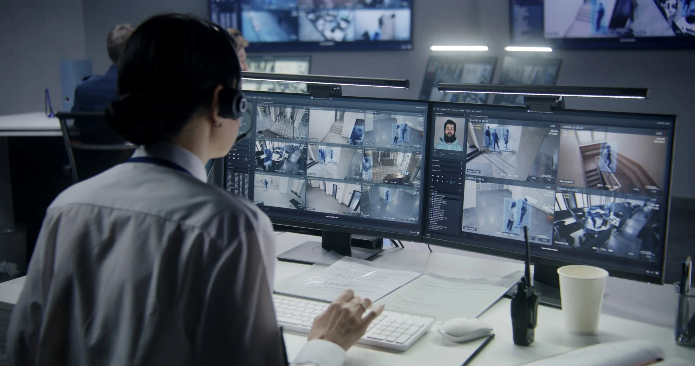 Security operator at a desk watching a wall of camera feeds, with a radio and coffee cup beside the keyboard