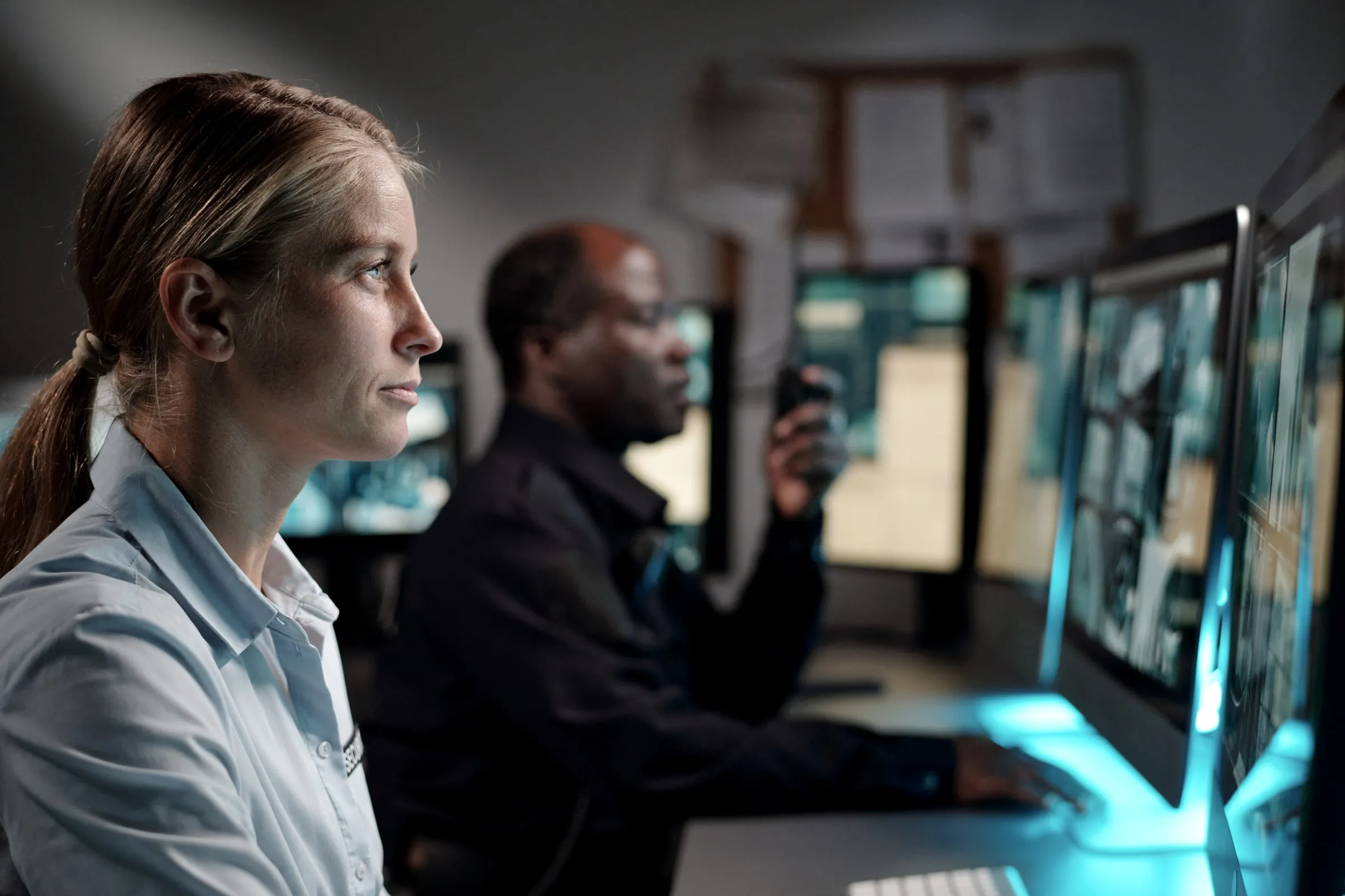 Woman in a light uniform shirt watching monitors in a dark room while a colleague behind her holds a radio