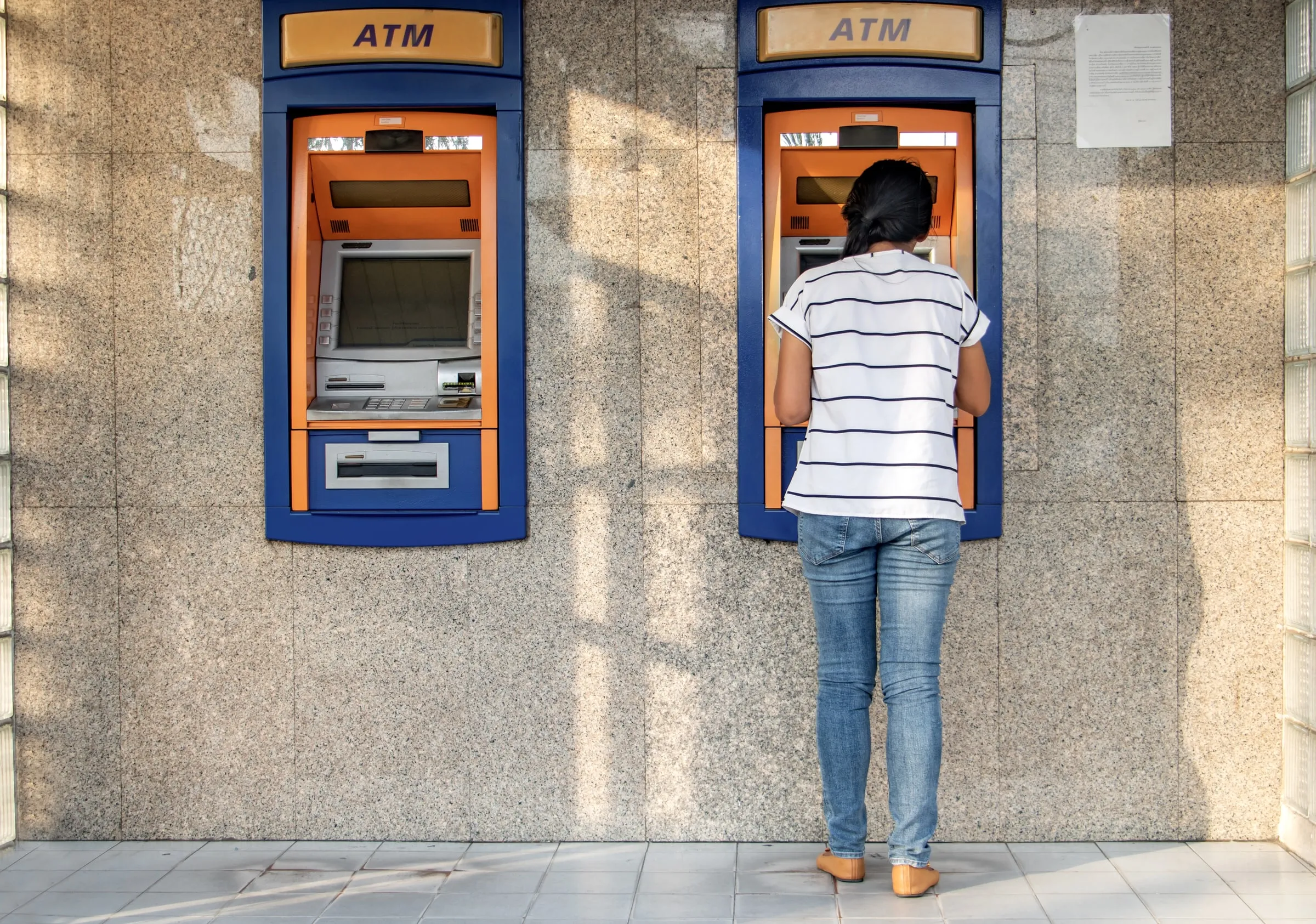 Woman using a walk-up ATM outdoors at a bank branch, seen from behind against a stone exterior wall