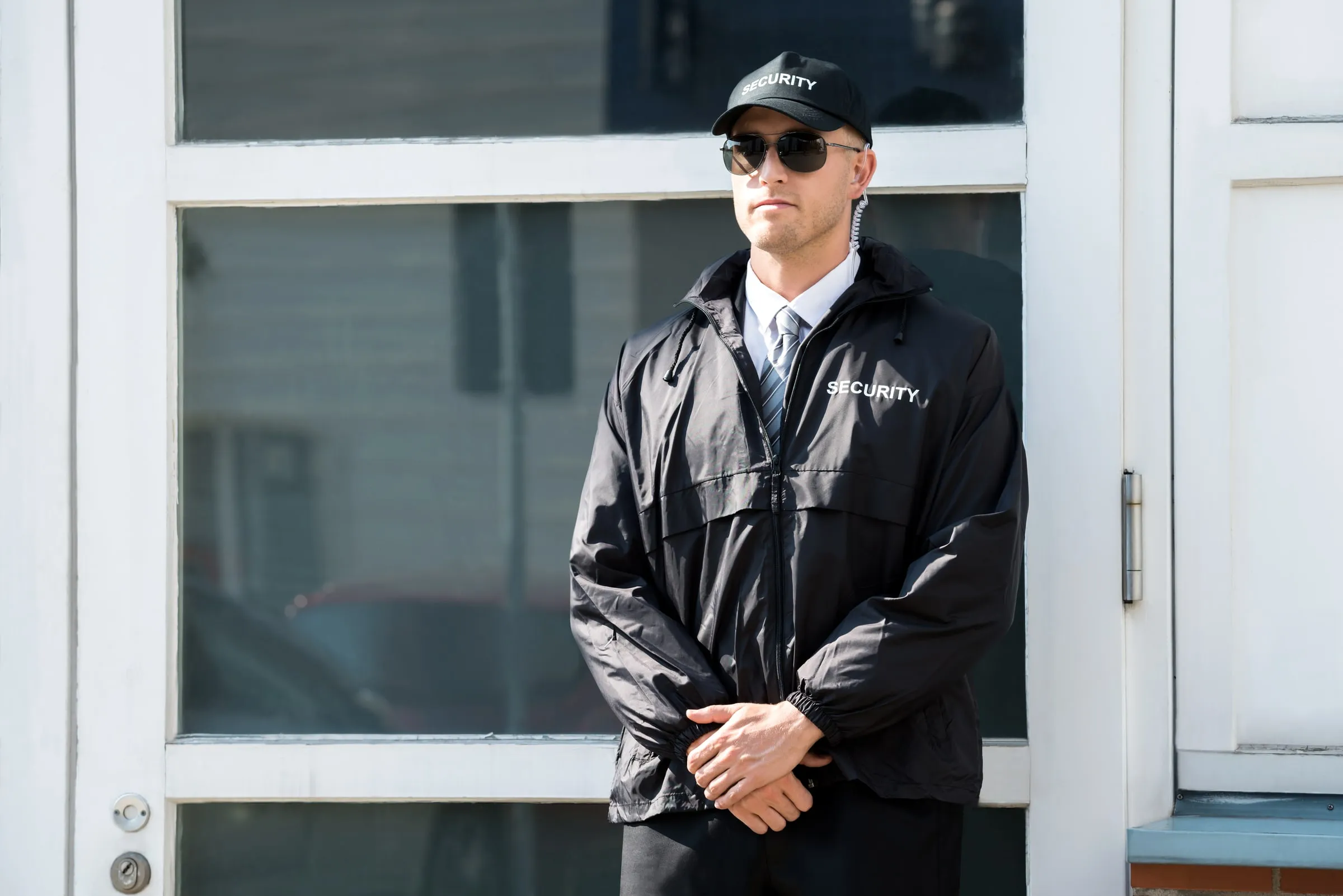 Security officer in a black jacket printed Security, cap and sunglasses standing with hands clasped in front of a white-framed glass entrance