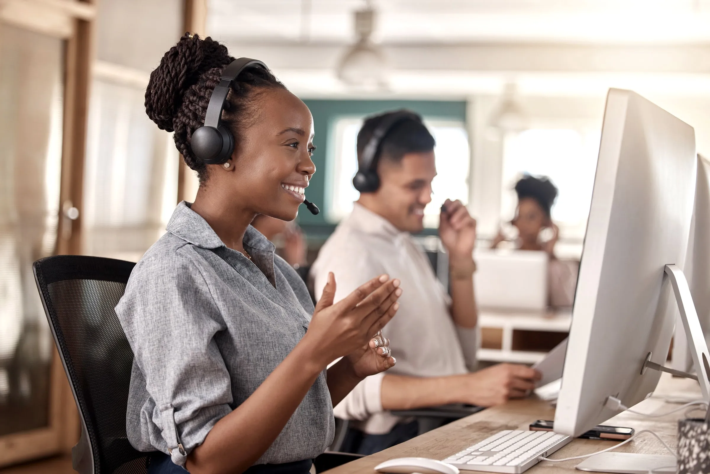 Woman wearing a headset smiling and clapping at a call center desk with colleagues working at monitors behind her