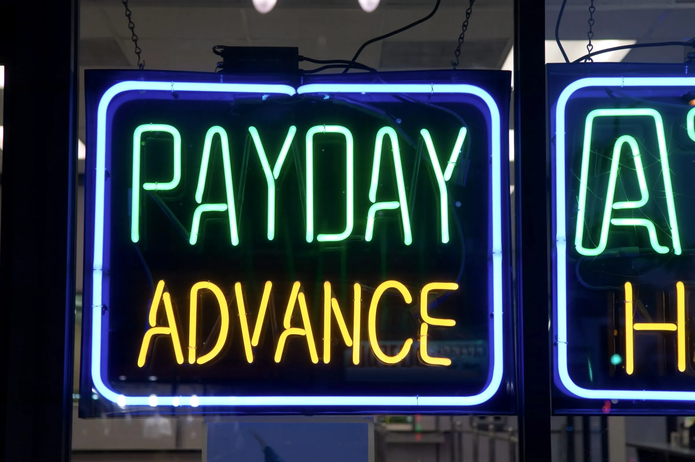 Illuminated neon storefront sign reading Payday Advance in green and yellow lettering inside a blue rectangle, seen through a shop window