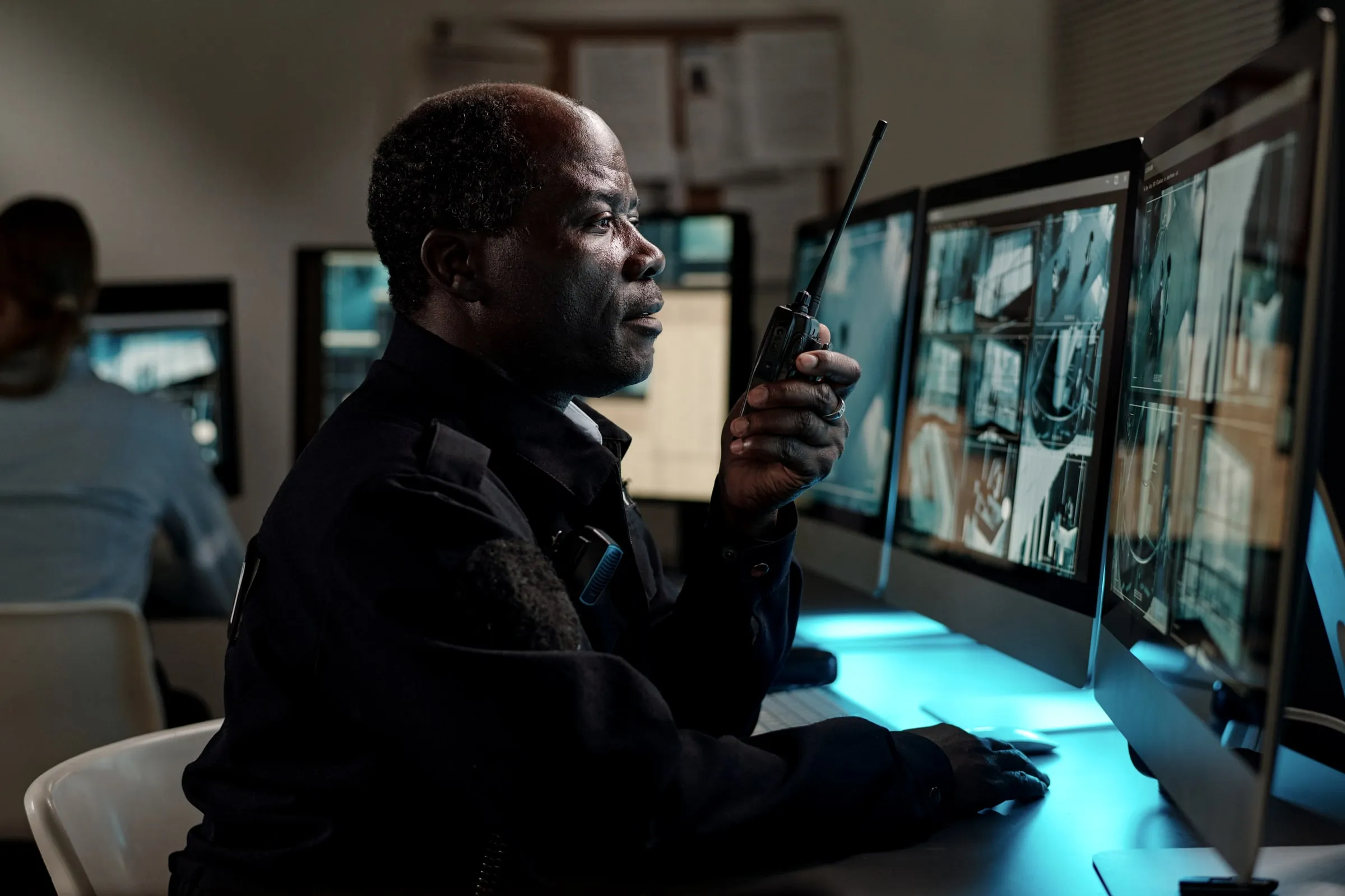 Security officer photographed from behind with hands clasped, wearing a duty belt with a radio, baton and handcuffs, against a plain sky background