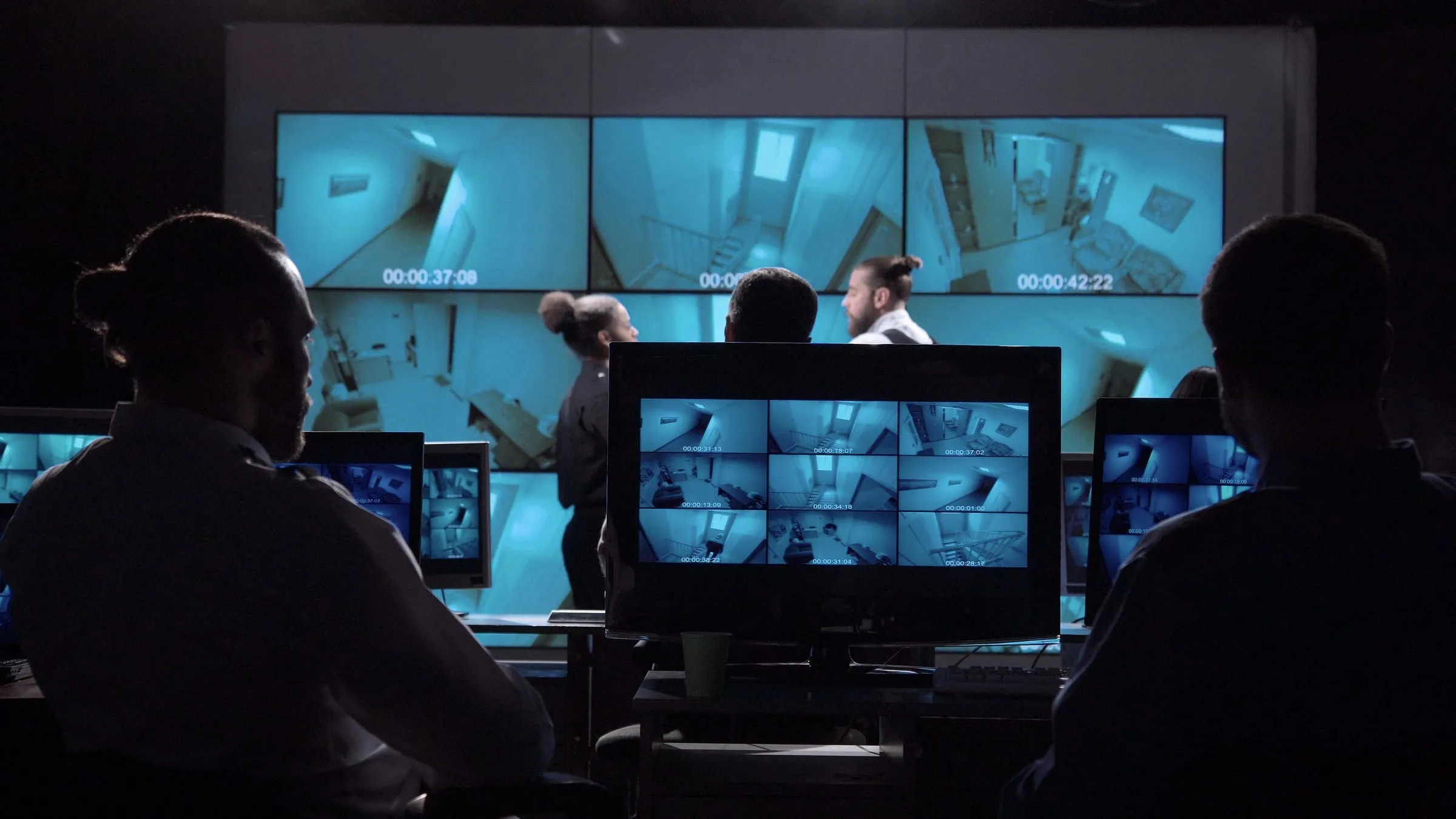 Three colleagues in a dark security control room reviewing a wide bank of monitors showing building interior camera feeds