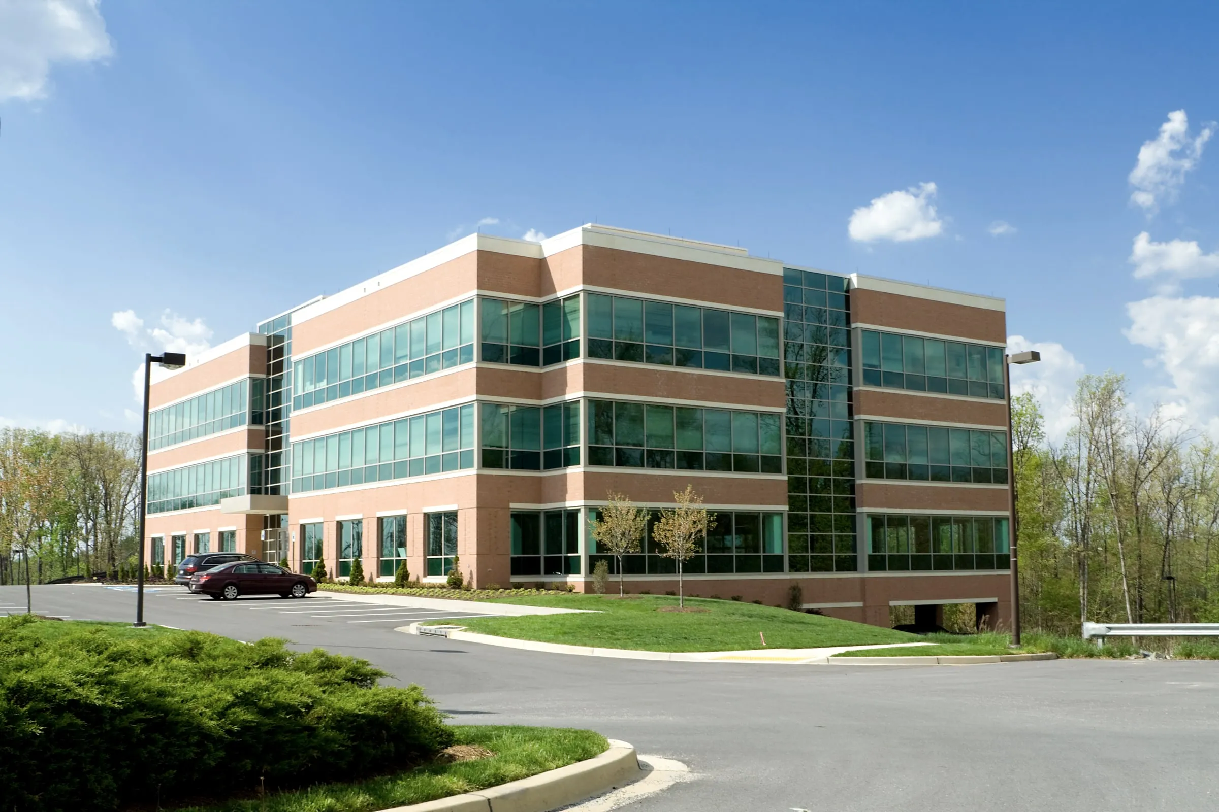 Four-story brick and glass corporate office building set among trees with a landscaped lawn and parking lot in front