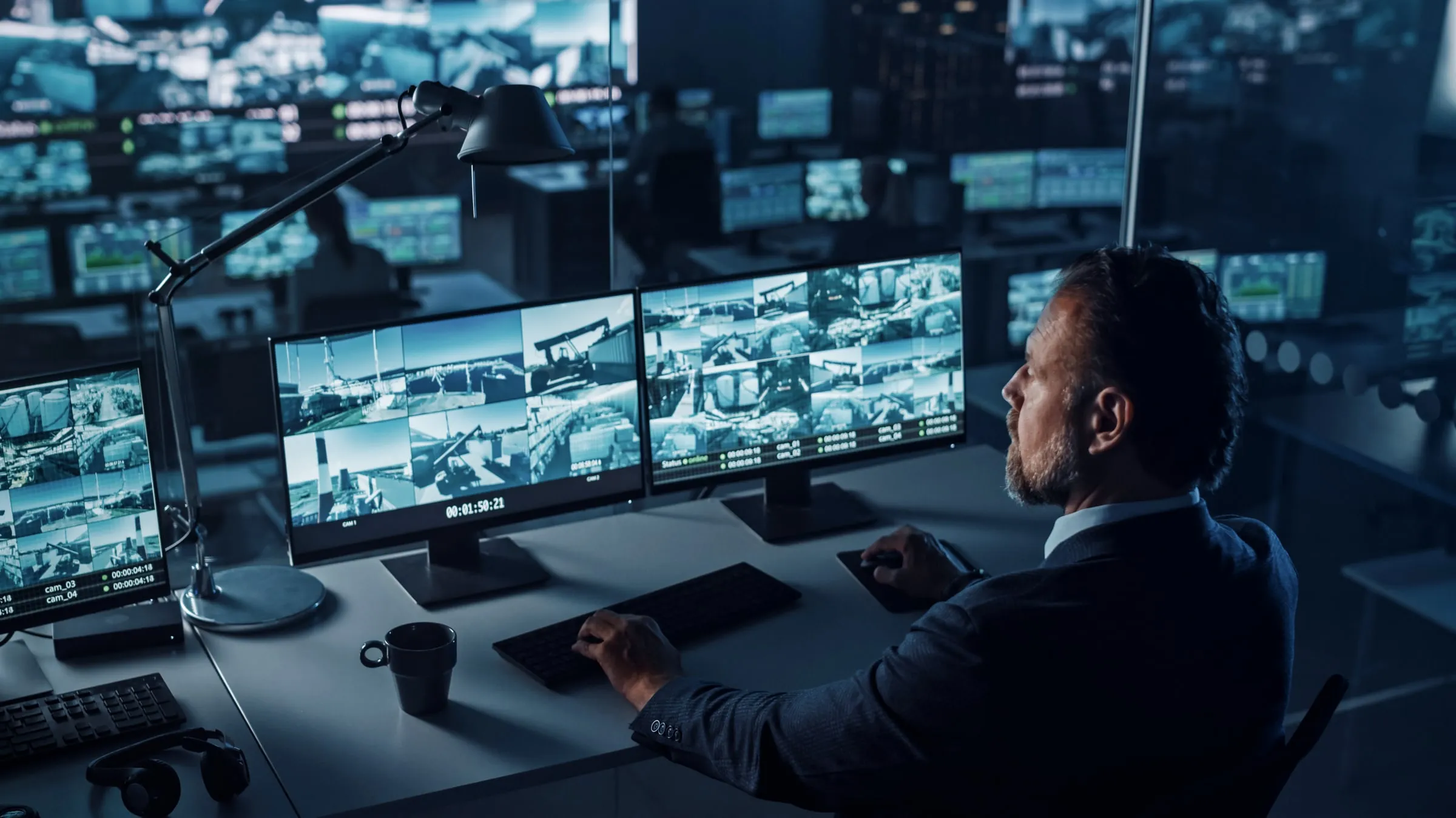 Security officer holding a radio while reviewing a bank of camera monitors showing corridor and dock footage in a dark control room