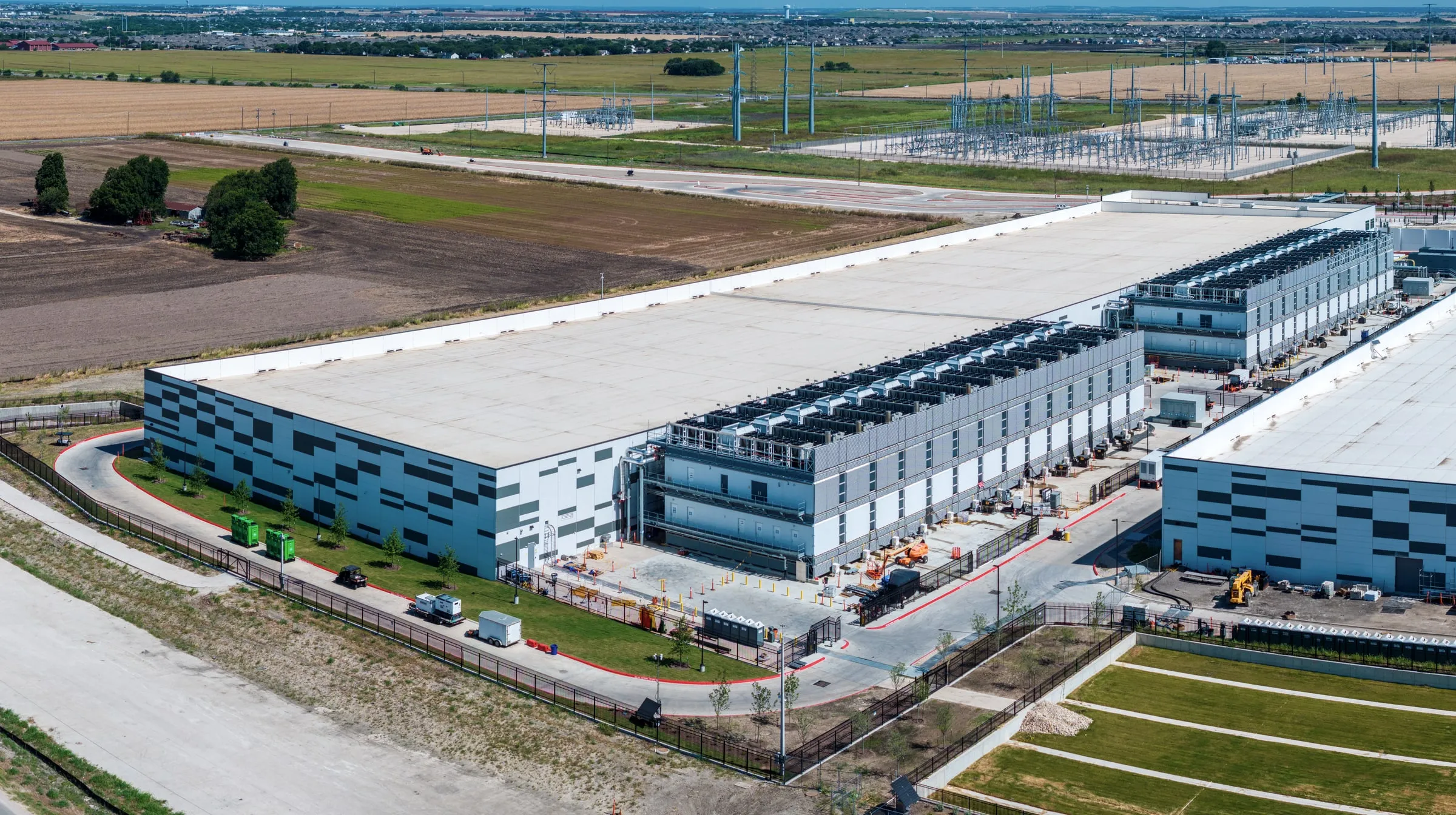 Aerial view of a large data center campus with server halls, rooftop cooling units, a substation and surrounding farmland
