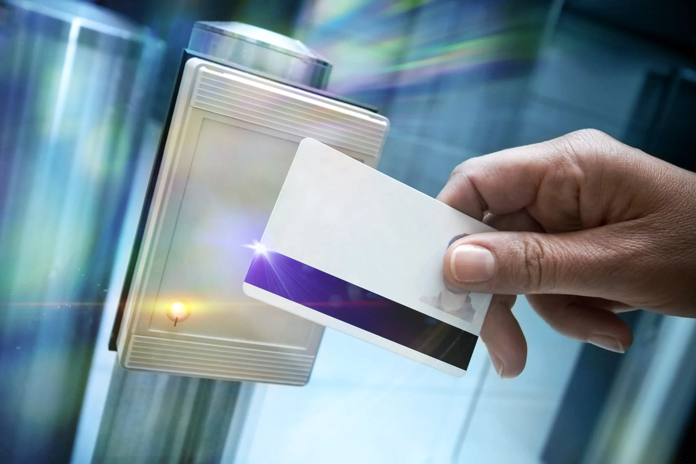 Hand holding a photo ID access card up to a wall-mounted card reader beside a glass door, with a small orange light indicator lit