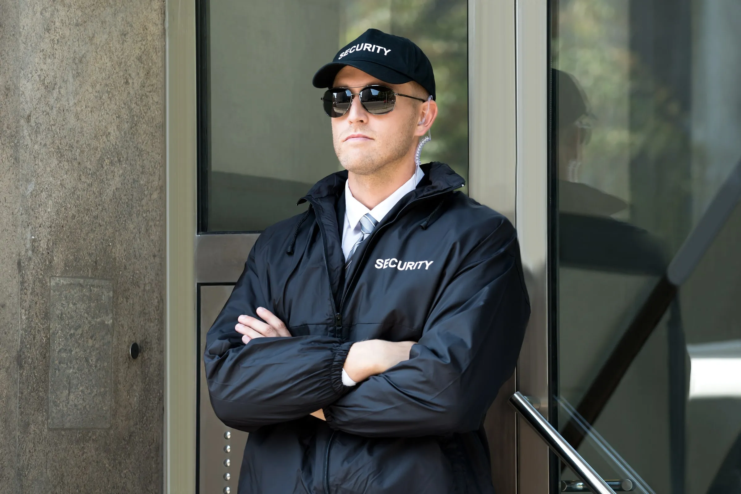 Security officer in a dark jacket printed Security, cap and sunglasses standing with arms crossed against a stone wall beside a glass entrance