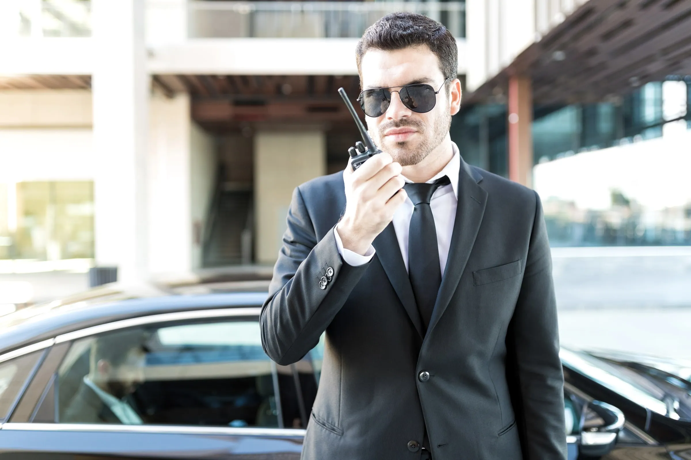 Man in a suit and aviator sunglasses speaking into a handheld radio outside a glass-fronted building, with a car and a colleague behind him