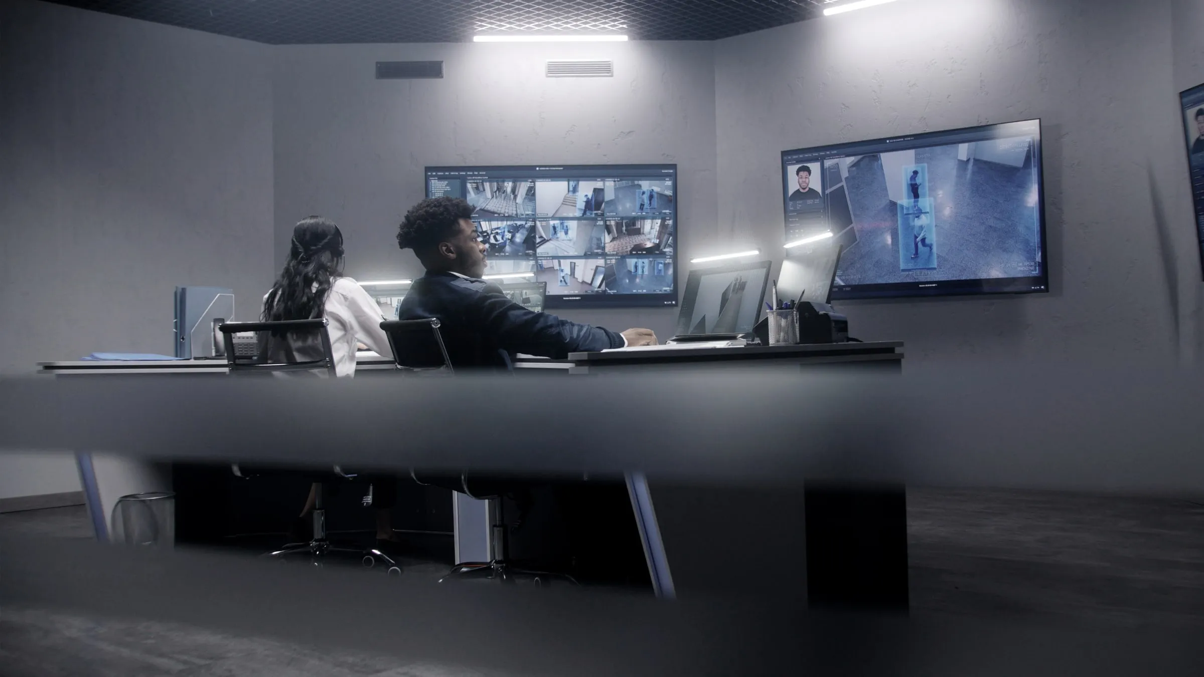 Two officers at a desk reviewing a wall of monitors showing hallway camera feeds and a visitor's photo pulled up on screen