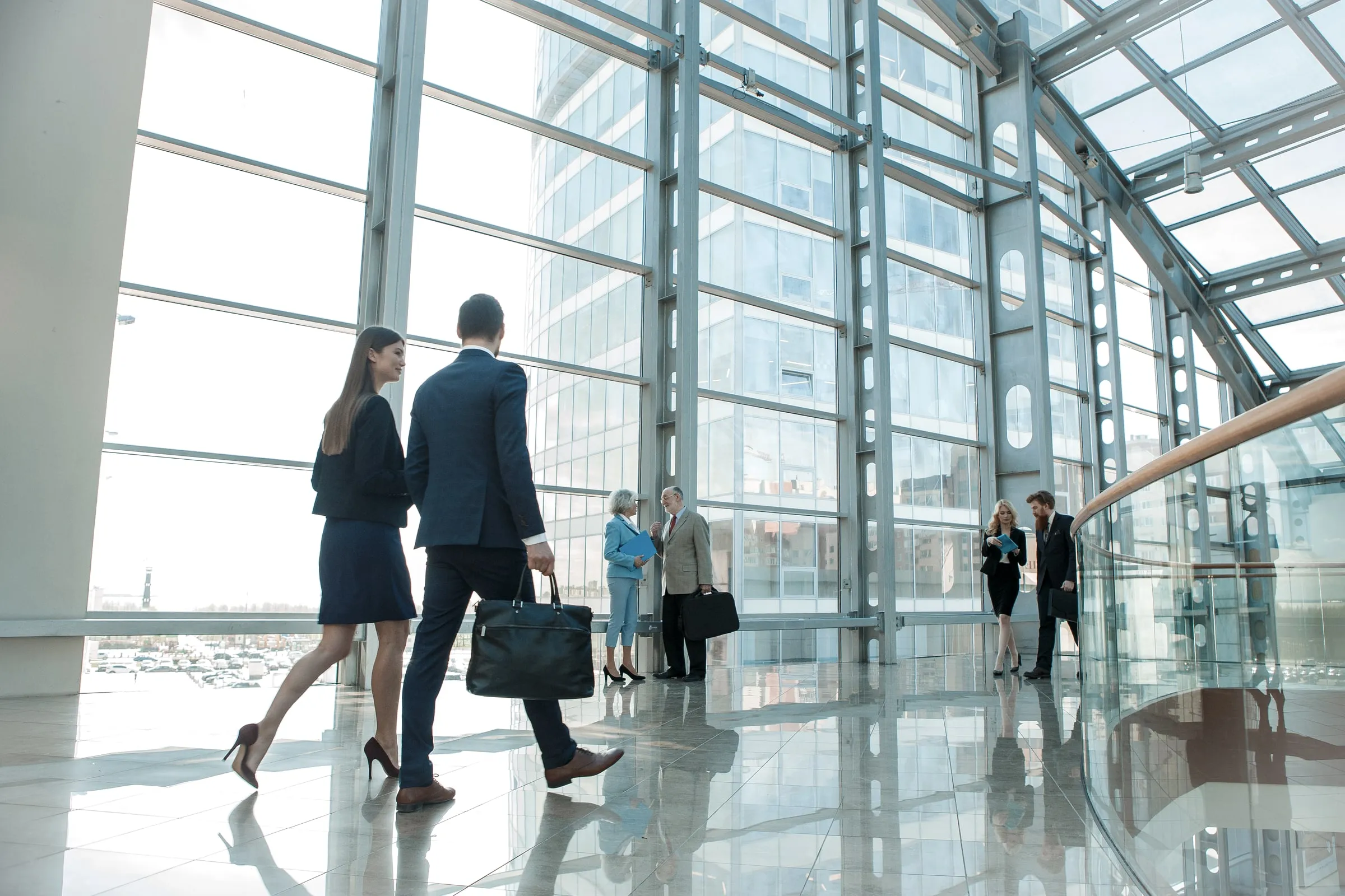 Two office workers walking through a glass-walled lobby atrium as colleagues meet near the windows and a curved stairwell railing