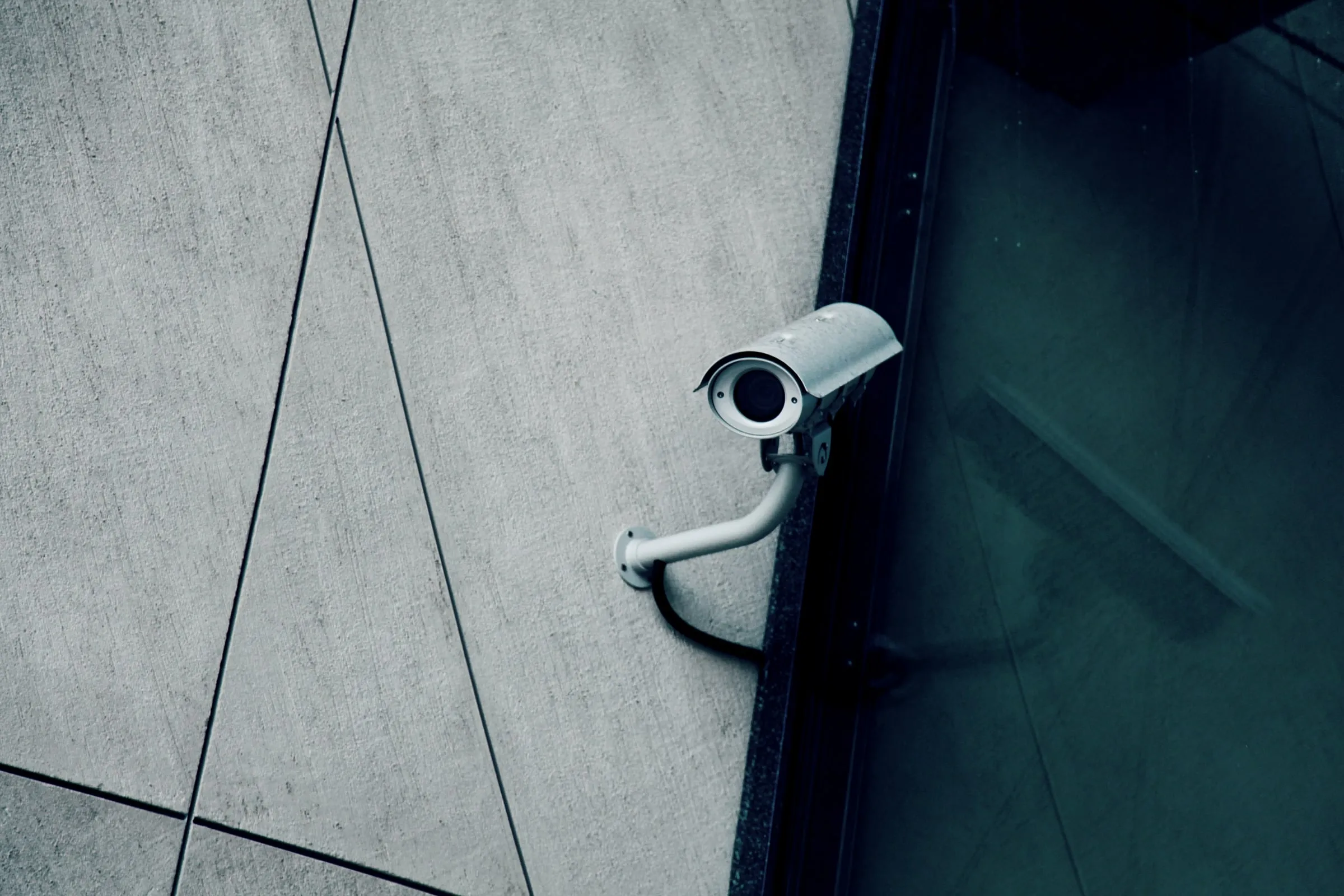 Security camera mounted on the corner of a building exterior beside a glass door, viewed from below at an angle