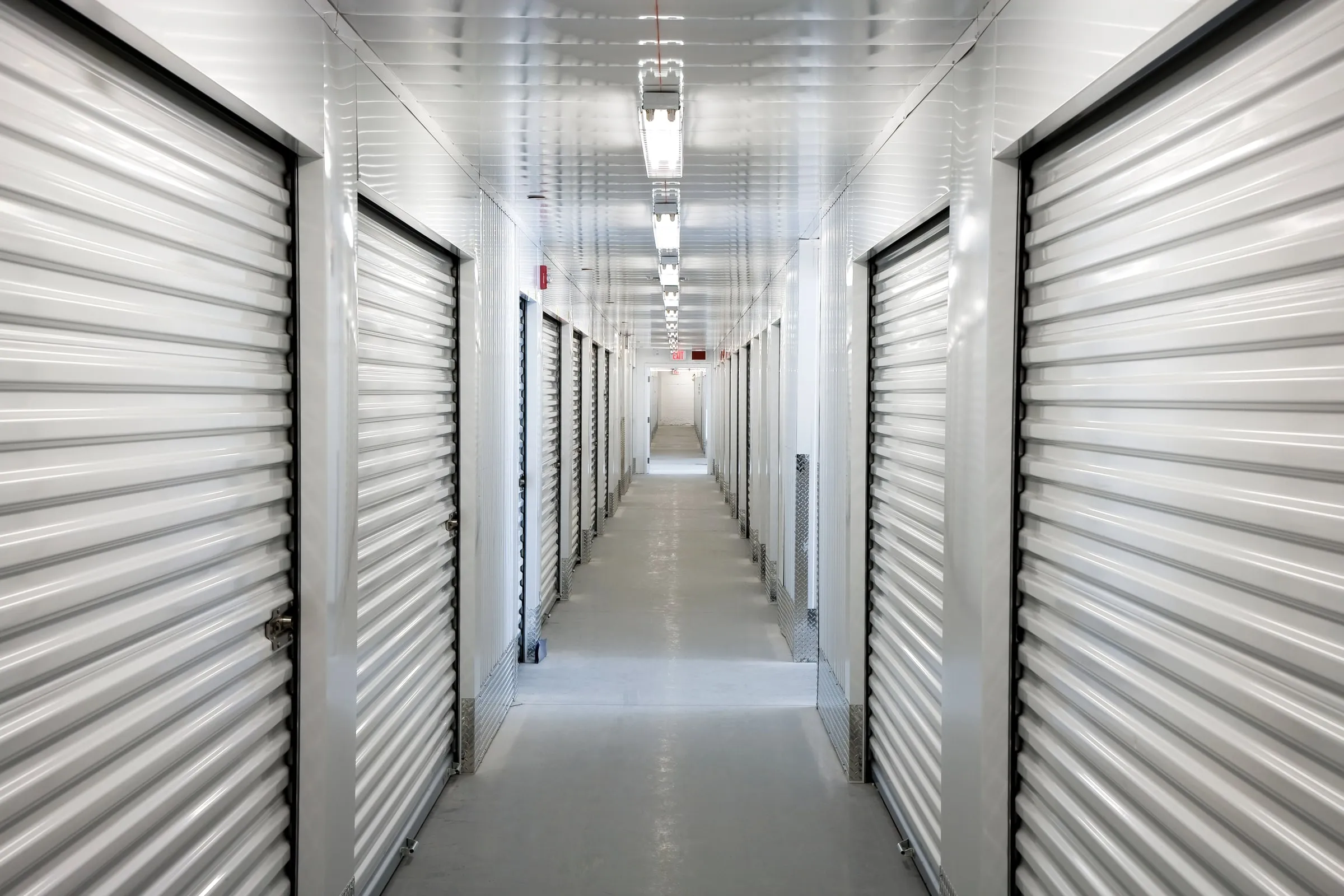 Long interior corridor of a self-storage facility lined with rows of white metal roll-up unit doors under fluorescent ceiling lights