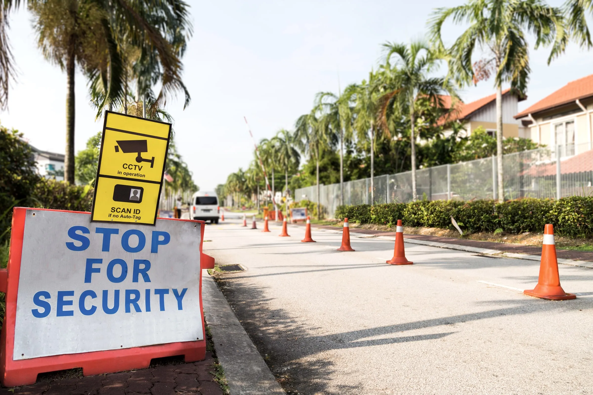 Portable Stop for Security barricade with CCTV and Scan ID signage and orange traffic cones along a palm-lined access road leading to a gated entrance