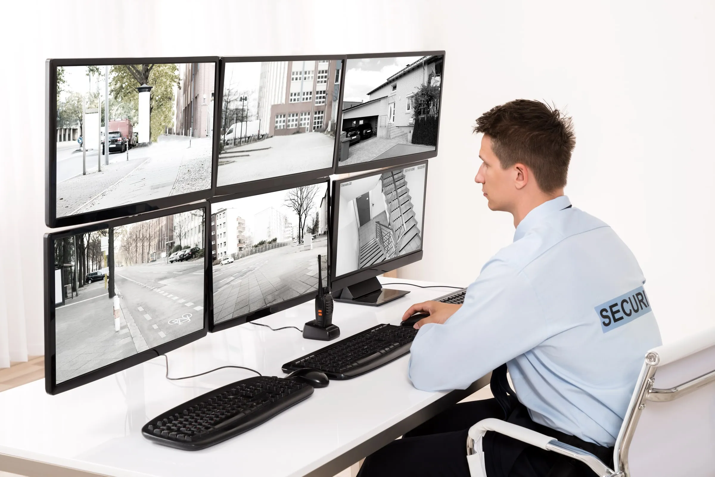 Security officer in a light blue uniform shirt seated at a desk watching six camera monitors that show streets and building entrances