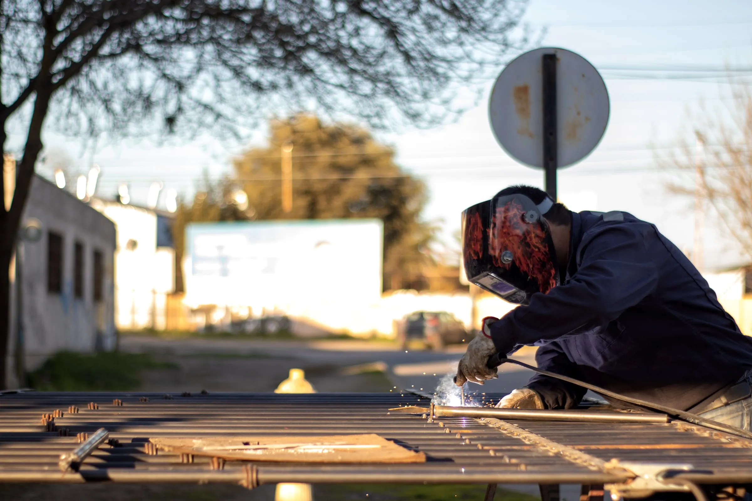Welder in a dark coverall and a flame-painted helmet welding a steel grate outdoors in low afternoon sun, sparks flying beside a road sign