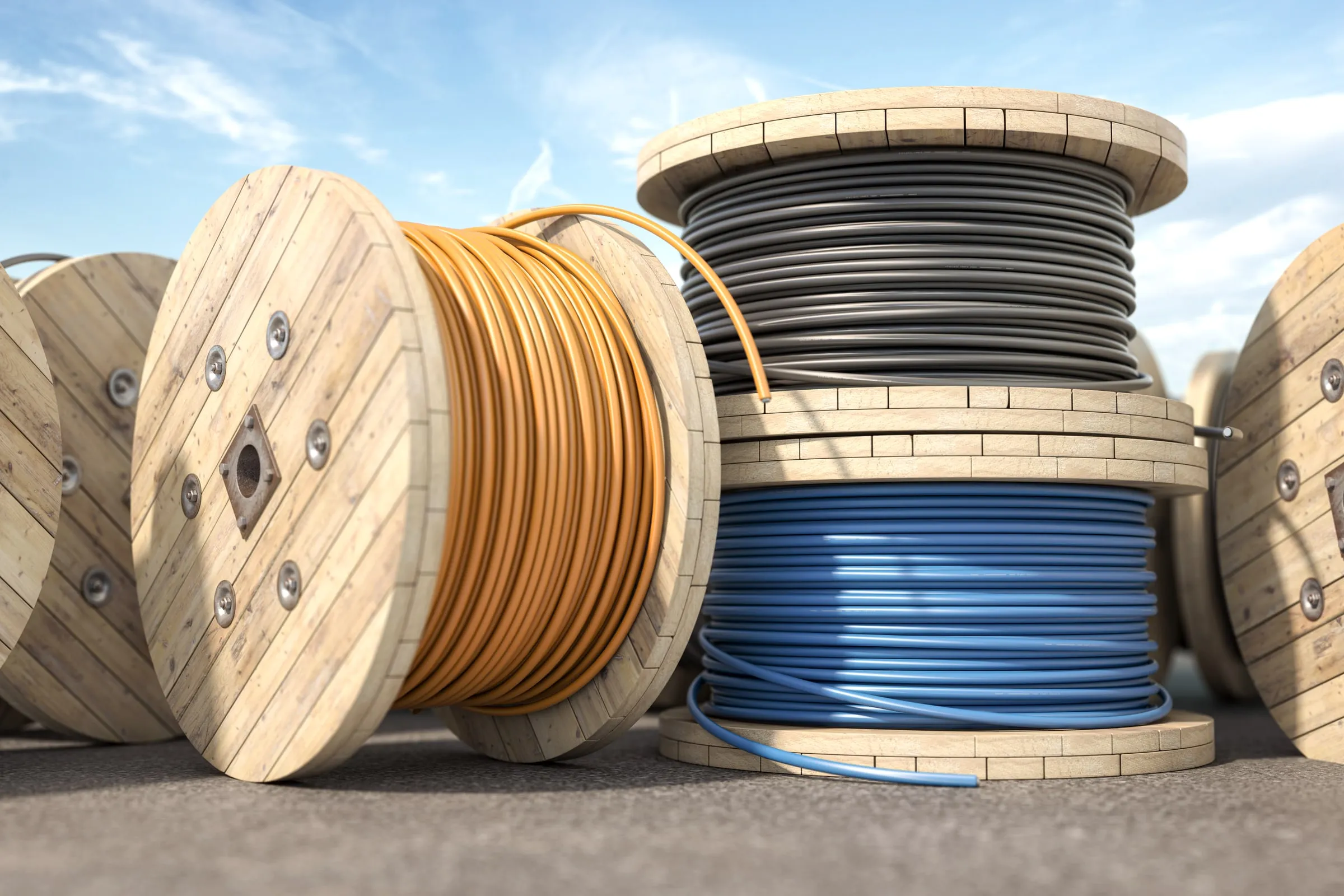 Large wooden cable reels wound with orange, blue and grey electrical cable standing on a paved yard under a blue sky