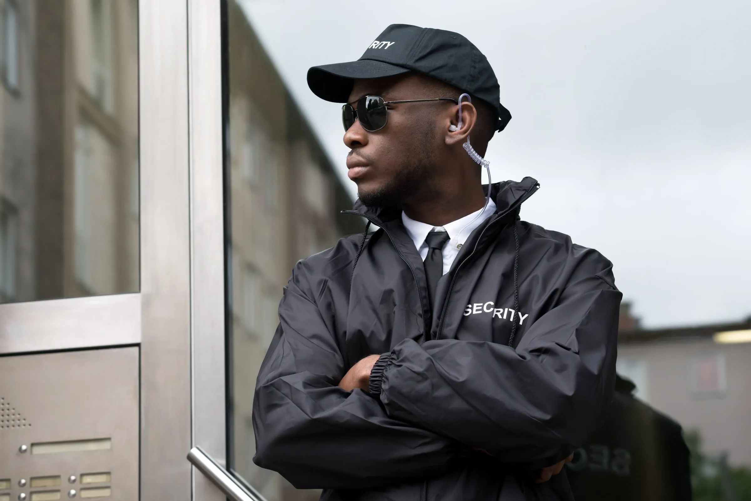 Security officer in a black jacket and cap with arms crossed standing beside a building entrance and intercom panel, looking off to the side
