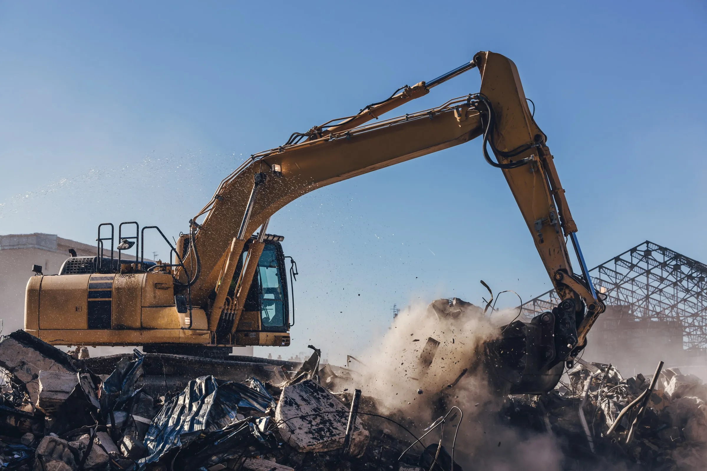 Yellow excavator with a demolition grapple tearing into a pile of debris and dust with a partially collapsed steel-framed structure visible behind it