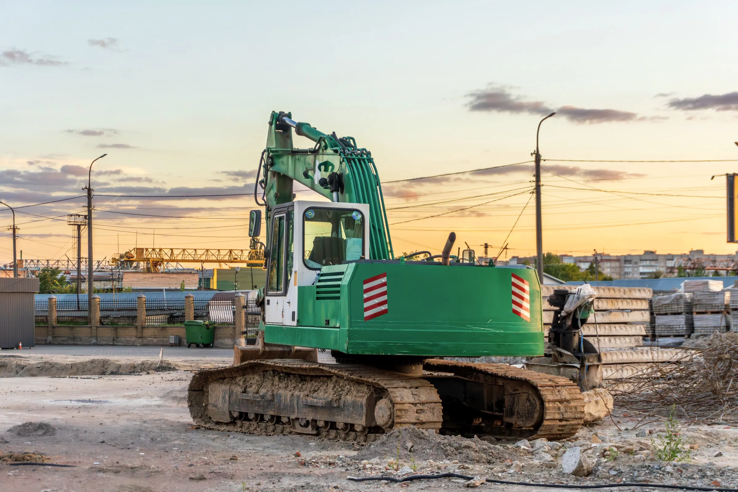 Green tracked excavator with hazard-striped panels parked at a construction yard at dusk beside stacked precast concrete sections and a city skyline