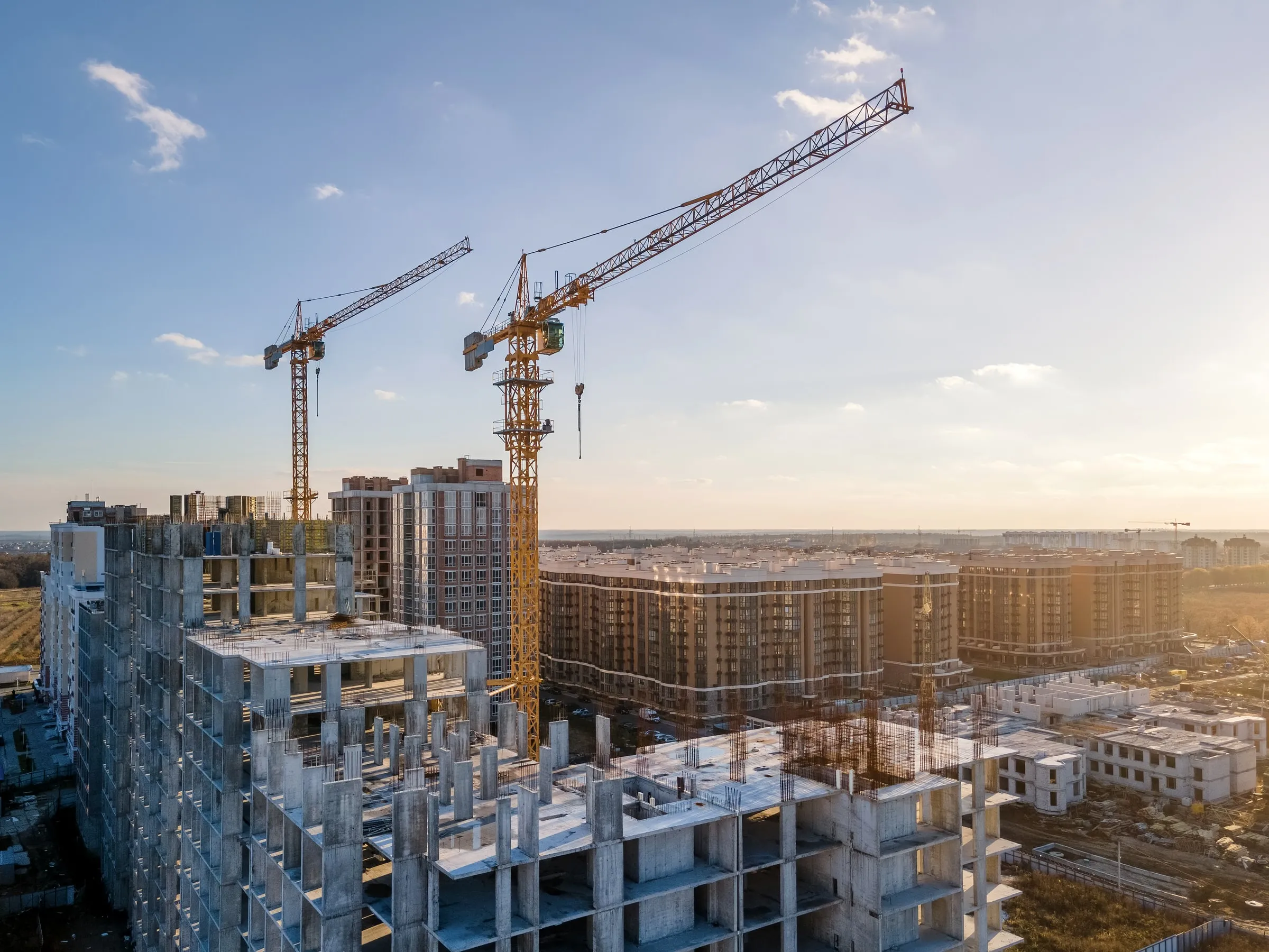 Tower cranes over a high-rise construction site at dusk, with a partly built concrete frame and city lights beyond