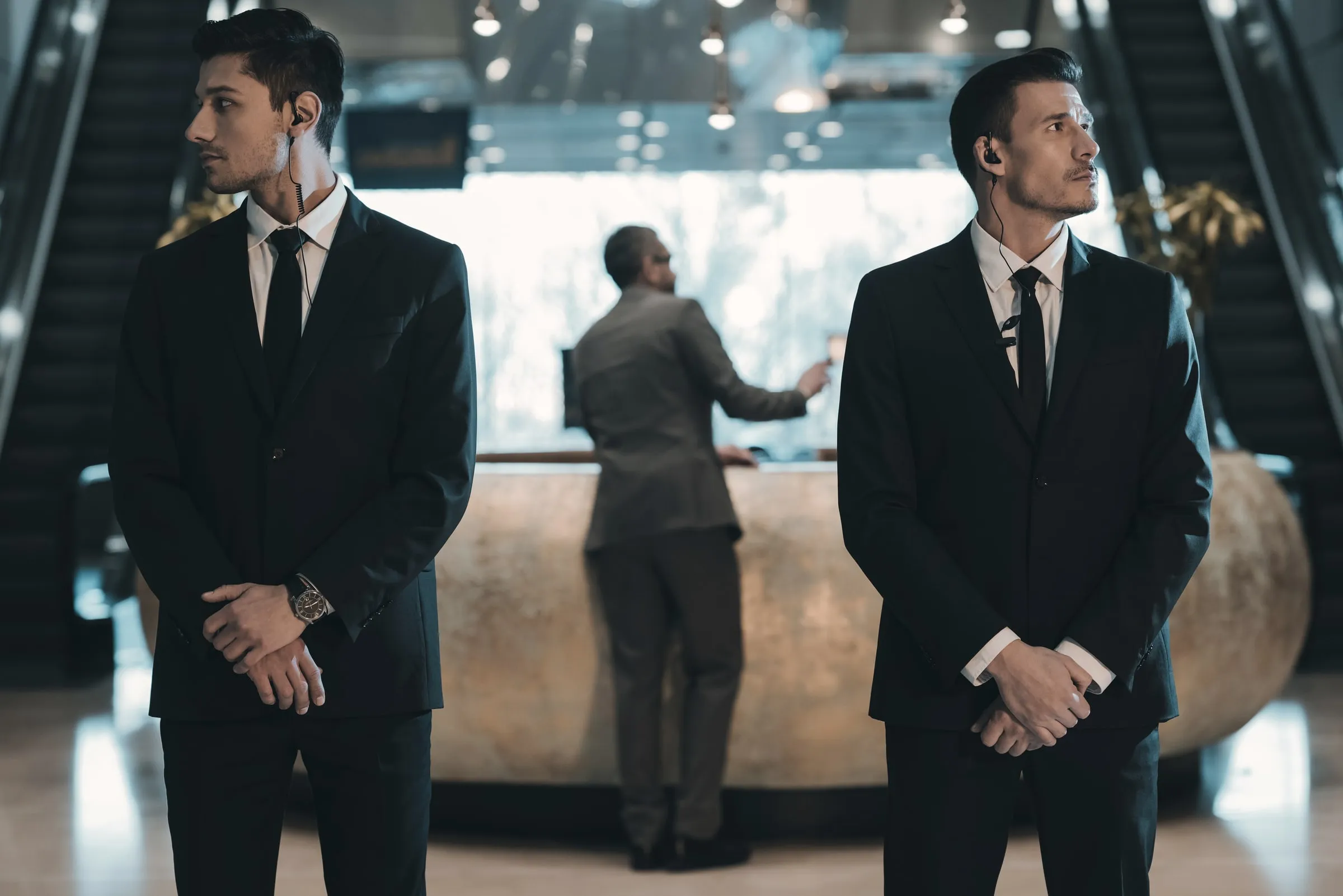 Two security officers in dark suits with earpieces standing watch in a marble lobby with an escalator, as a third man works at a reception counter behind them