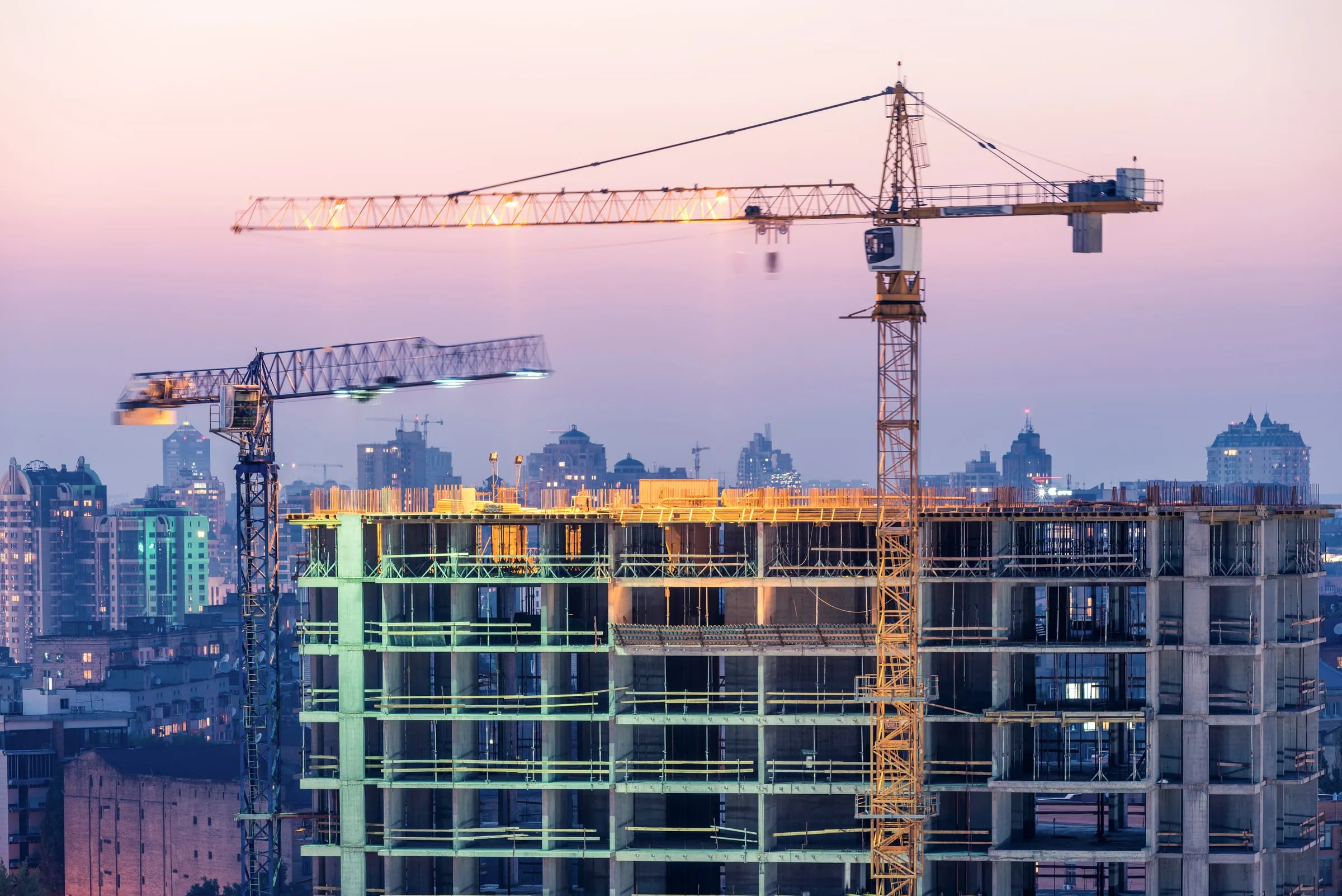 Two tower cranes beside a high-rise building under construction with exposed concrete floors, photographed at dusk against a pink and purple sky
