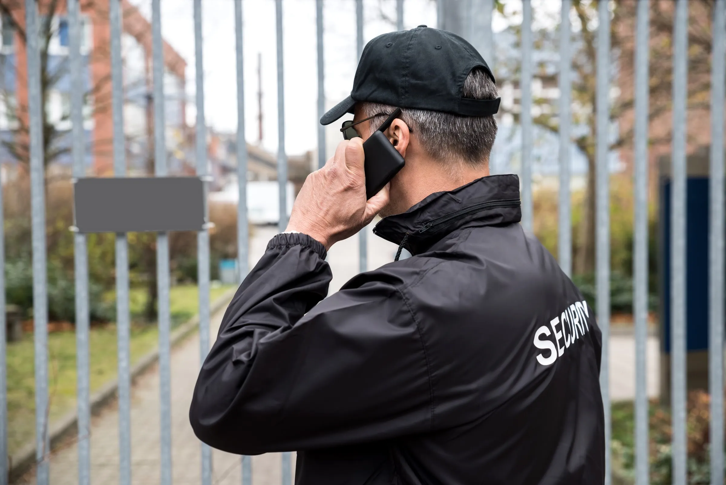 Security officer viewed from behind wearing a black jacket printed with Security, speaking on a cell phone in front of a metal picket fence with residential buildings beyond