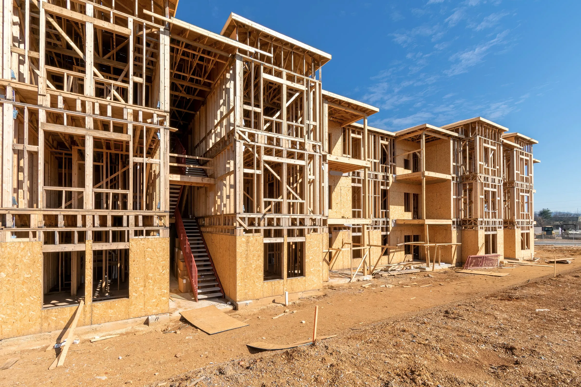 Row of wood-framed multi-unit apartment buildings under construction with exterior stairwells and multiple balcony openings, seen against a blue sky