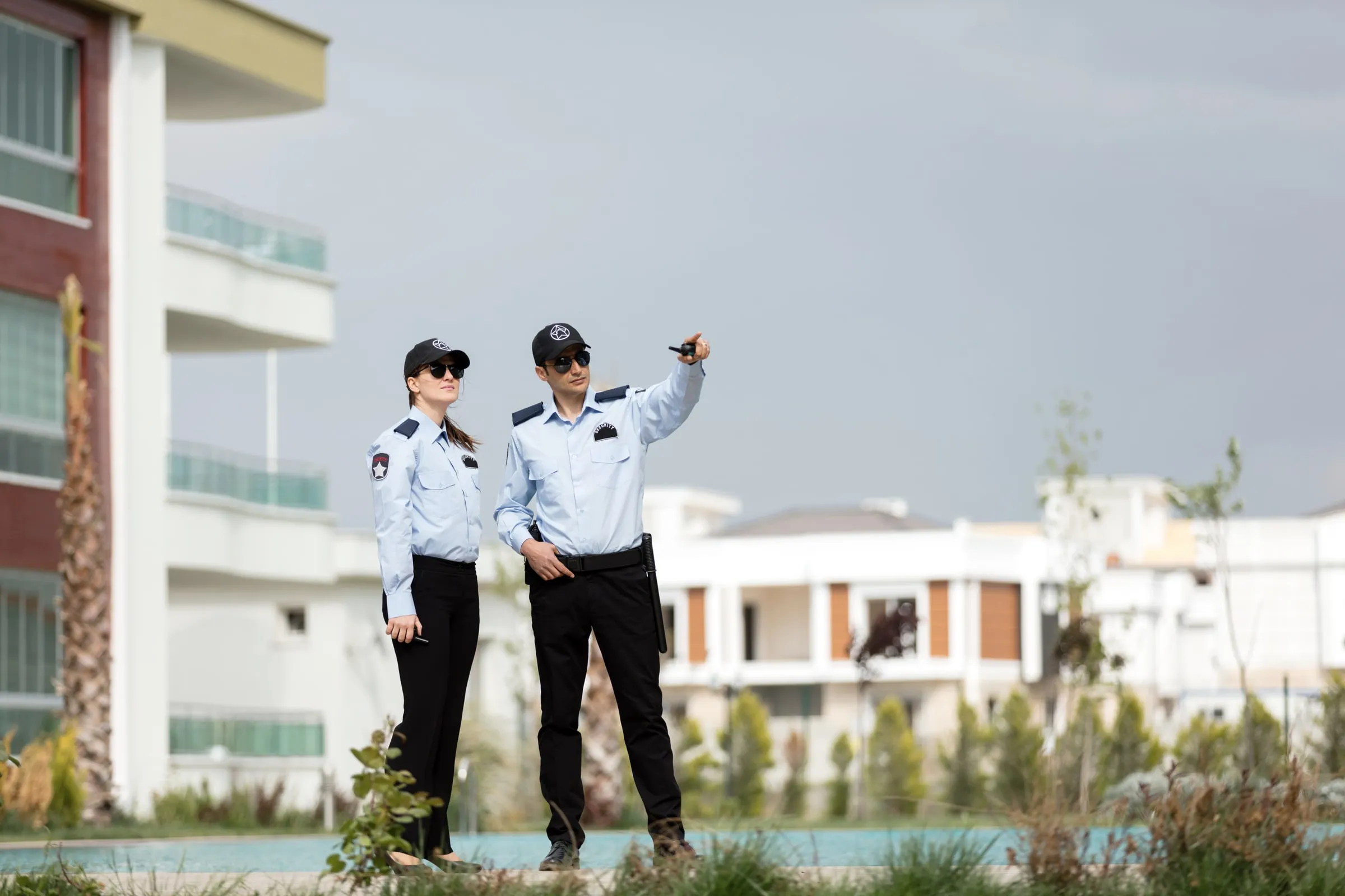 Two security officers in light blue uniform shirts and caps standing outdoors near a residential building, one pointing while holding a handheld radio