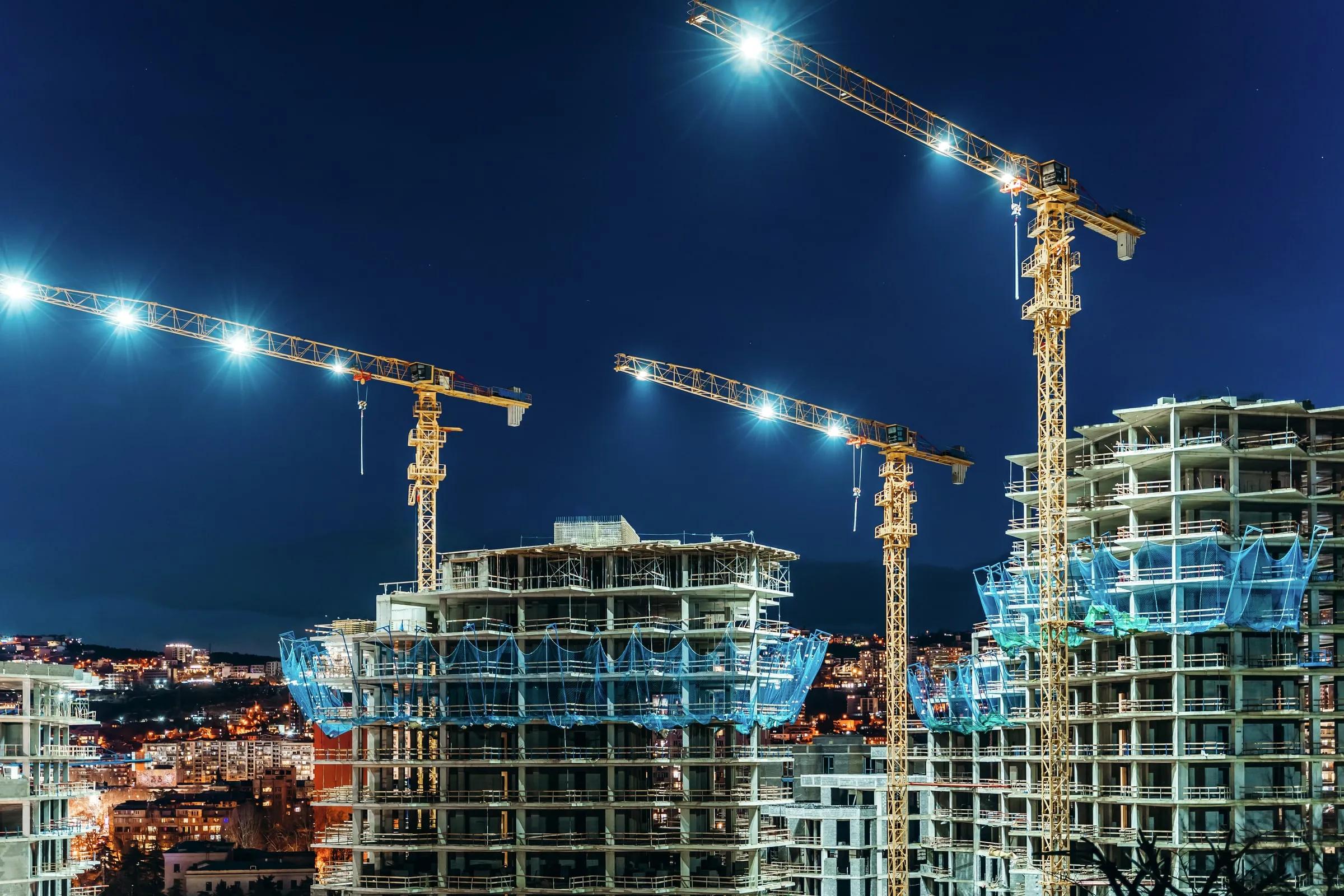 Two tower cranes lit against a night sky beside high-rise buildings under construction wrapped in blue safety netting, city lights below