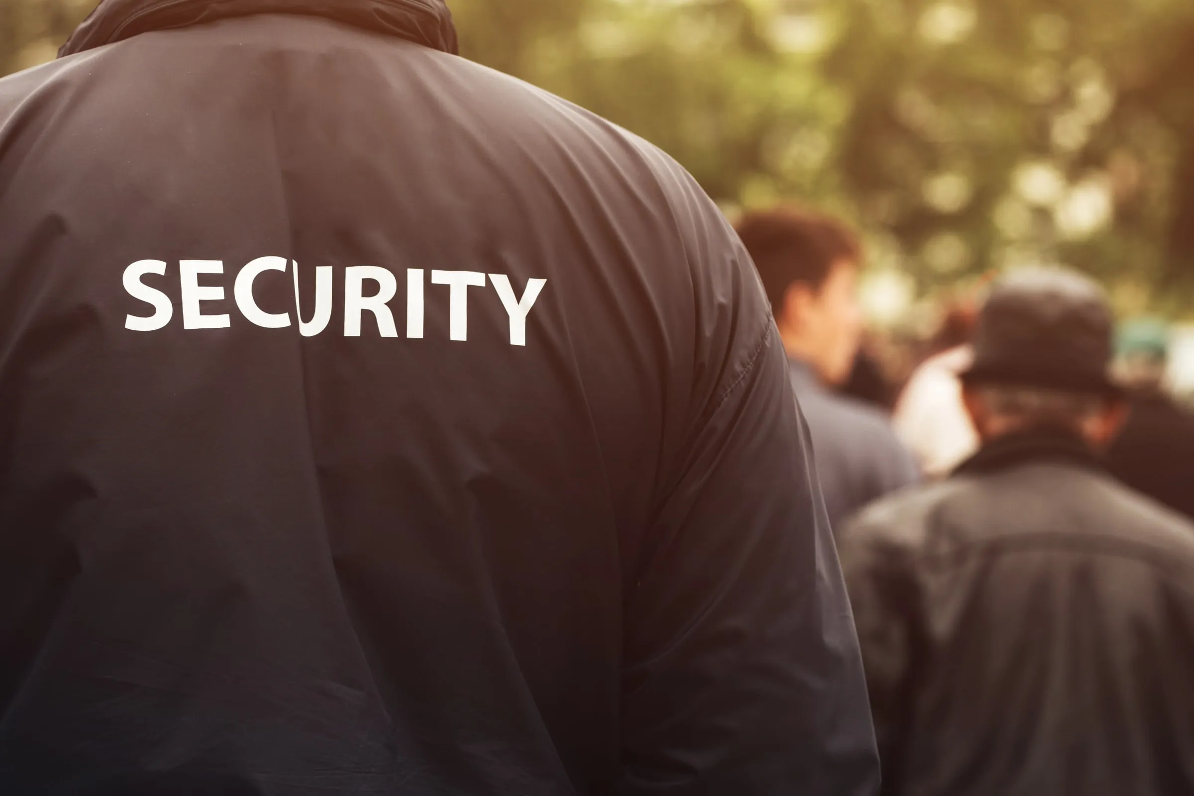 Back of a security officer in a dark jacket with SECURITY printed across the shoulders, facing a blurred group of people among trees