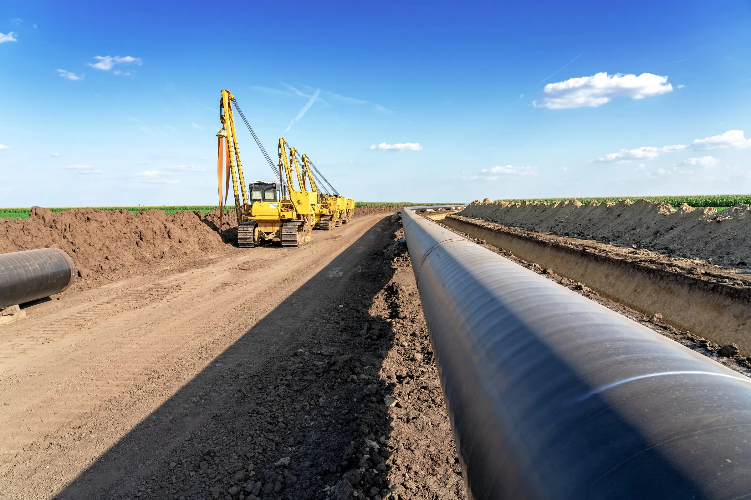 Long black pipeline laid beside an open trench and a dirt access road, with yellow pipelayer tractors parked in the distance under a blue sky