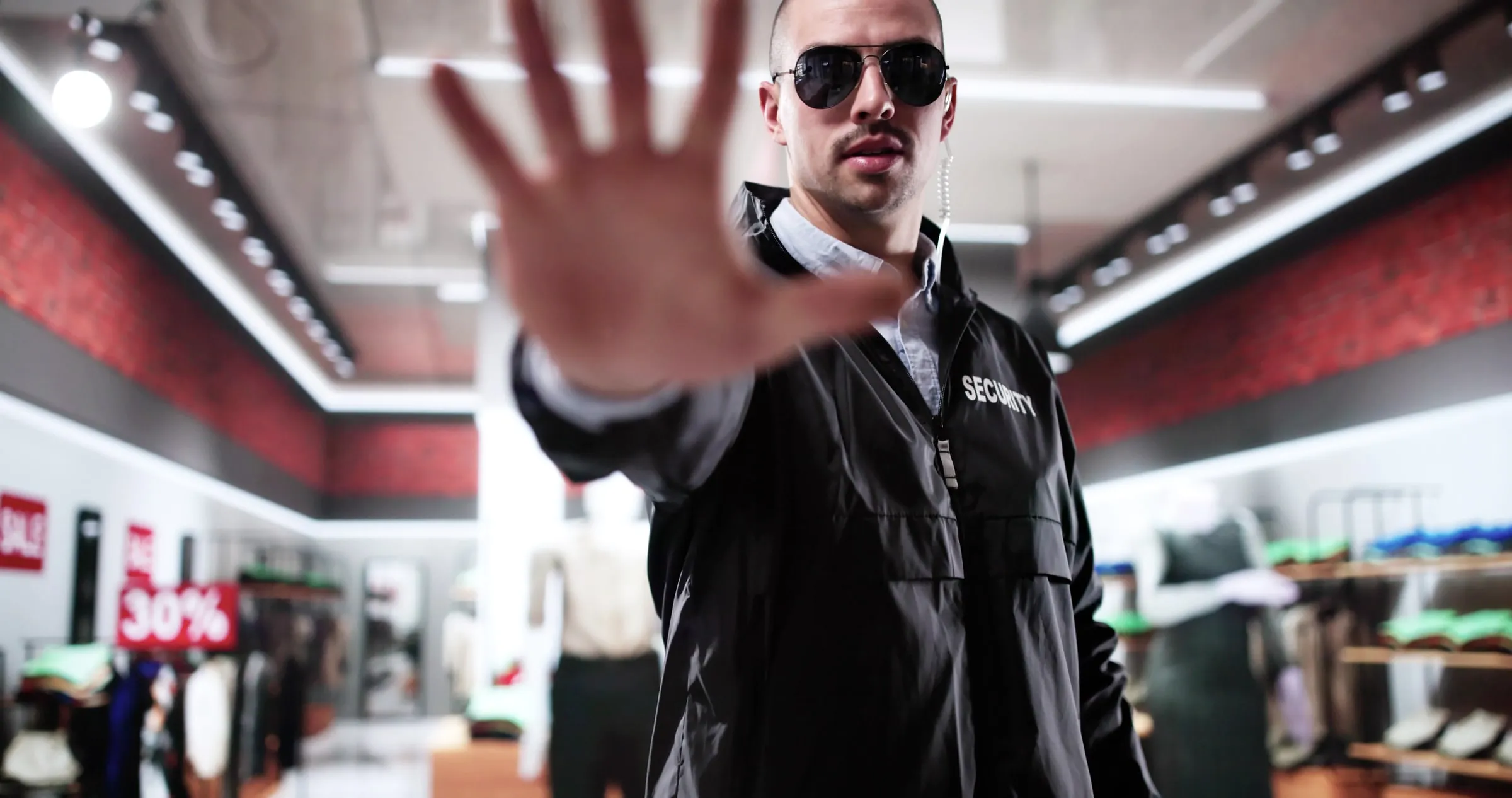 Security officer in a black jacket printed with Security raising a hand toward the camera while standing inside a retail clothing store with a sale sign