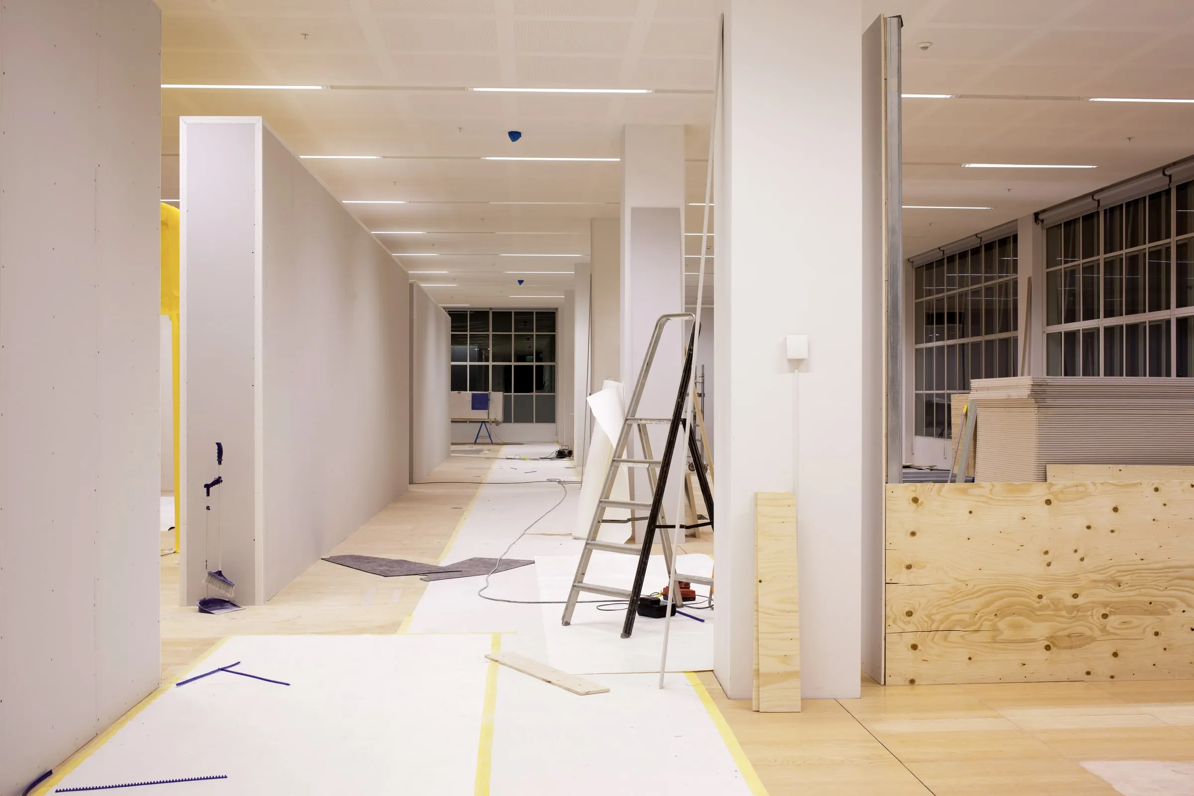 Office interior mid-renovation with exposed studs, a stepladder, plywood partitions and construction materials laid on a taped-off plywood floor
