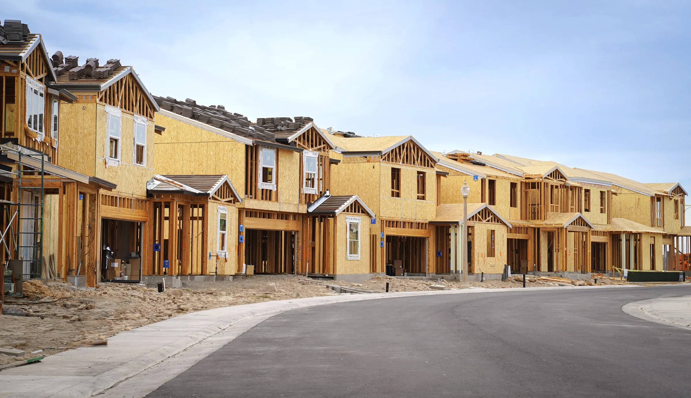 Row of wood-framed single-family homes under construction along a curving paved subdivision street, with roof tile stacked on one roof and framing exposed