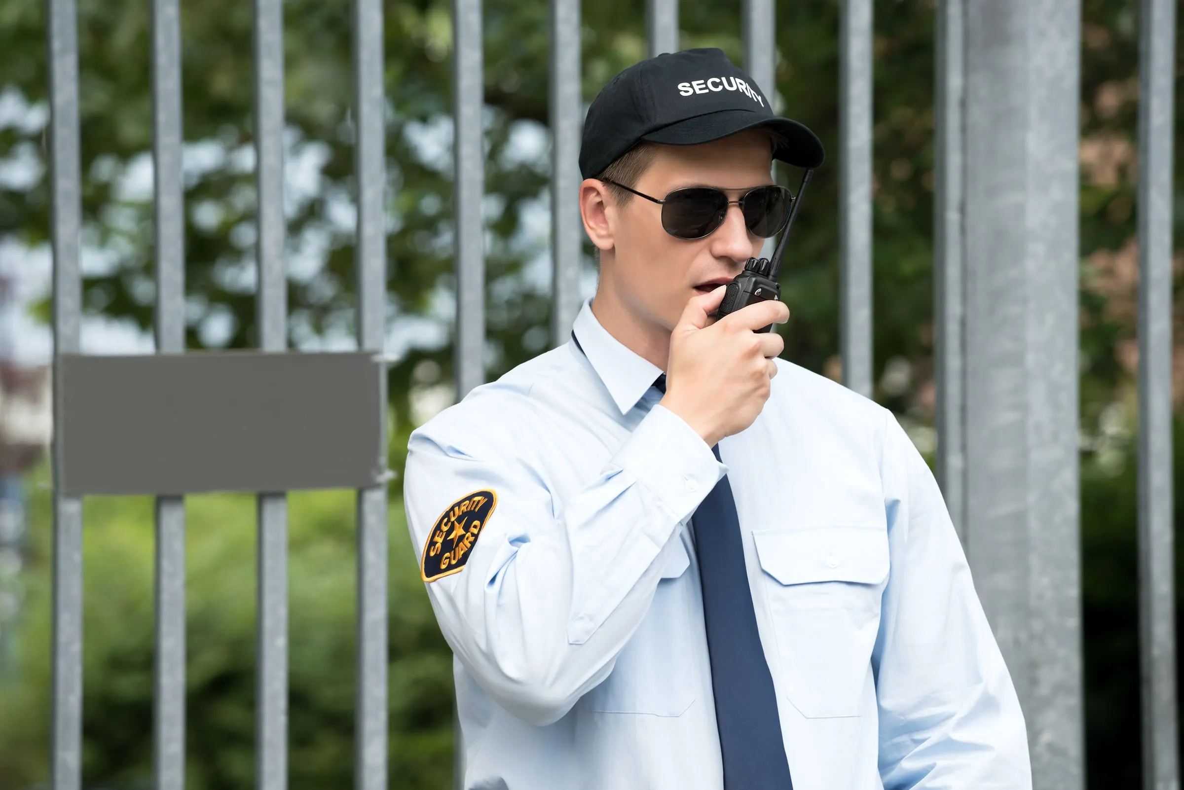 Security officer in a light blue uniform shirt with a Security Guard patch, cap and sunglasses speaking into a handheld radio in front of a metal fence