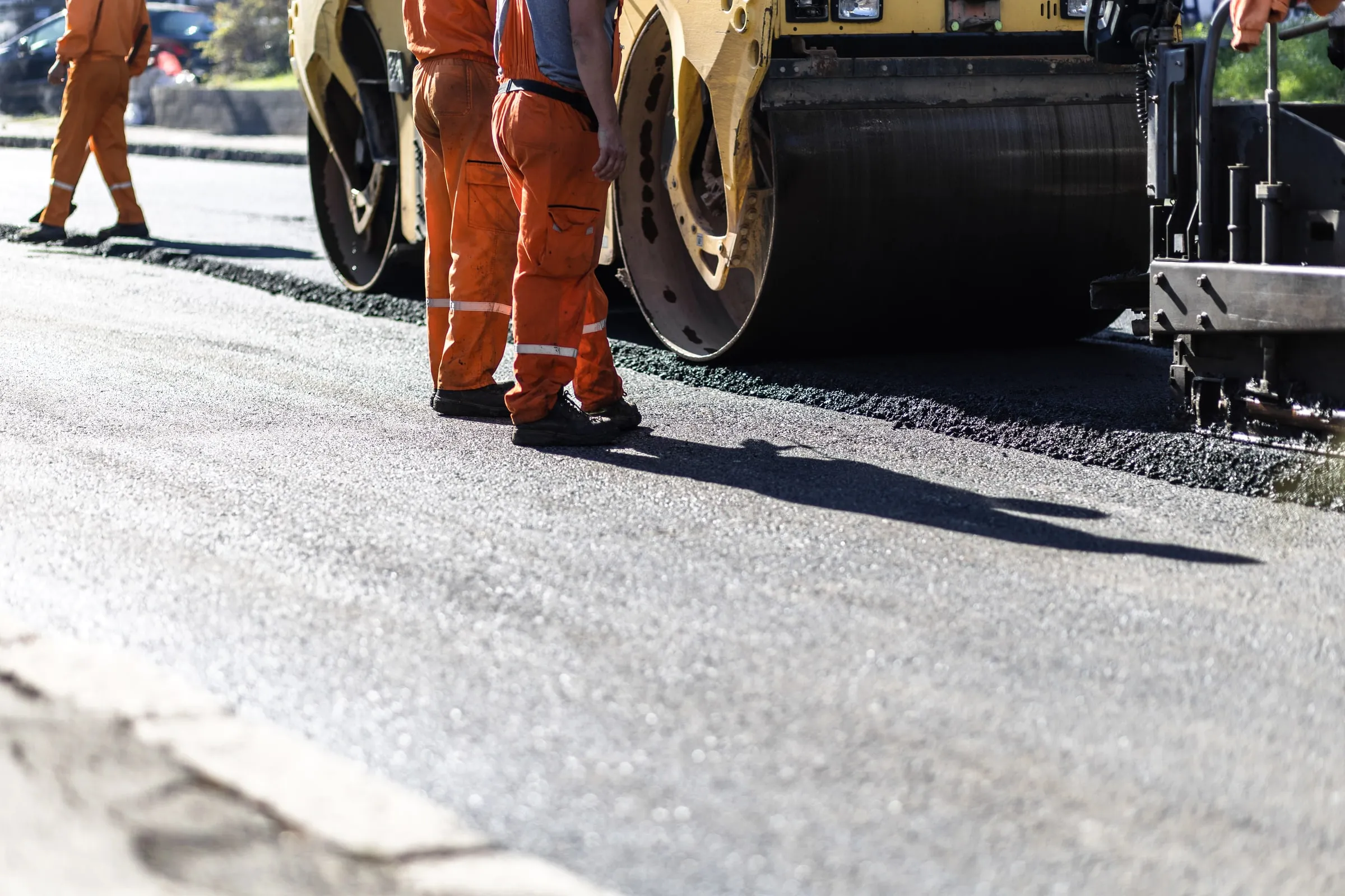 Two road crew workers in orange coveralls standing beside a yellow roller compactor and asphalt paver laying fresh blacktop on a street