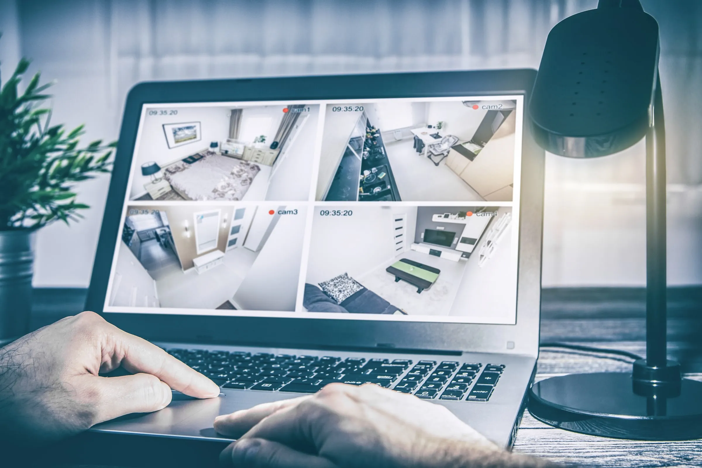 Person's hands on a laptop keyboard displaying four security camera feeds of a bedroom, kitchen, hallway and living room, beside a desk lamp