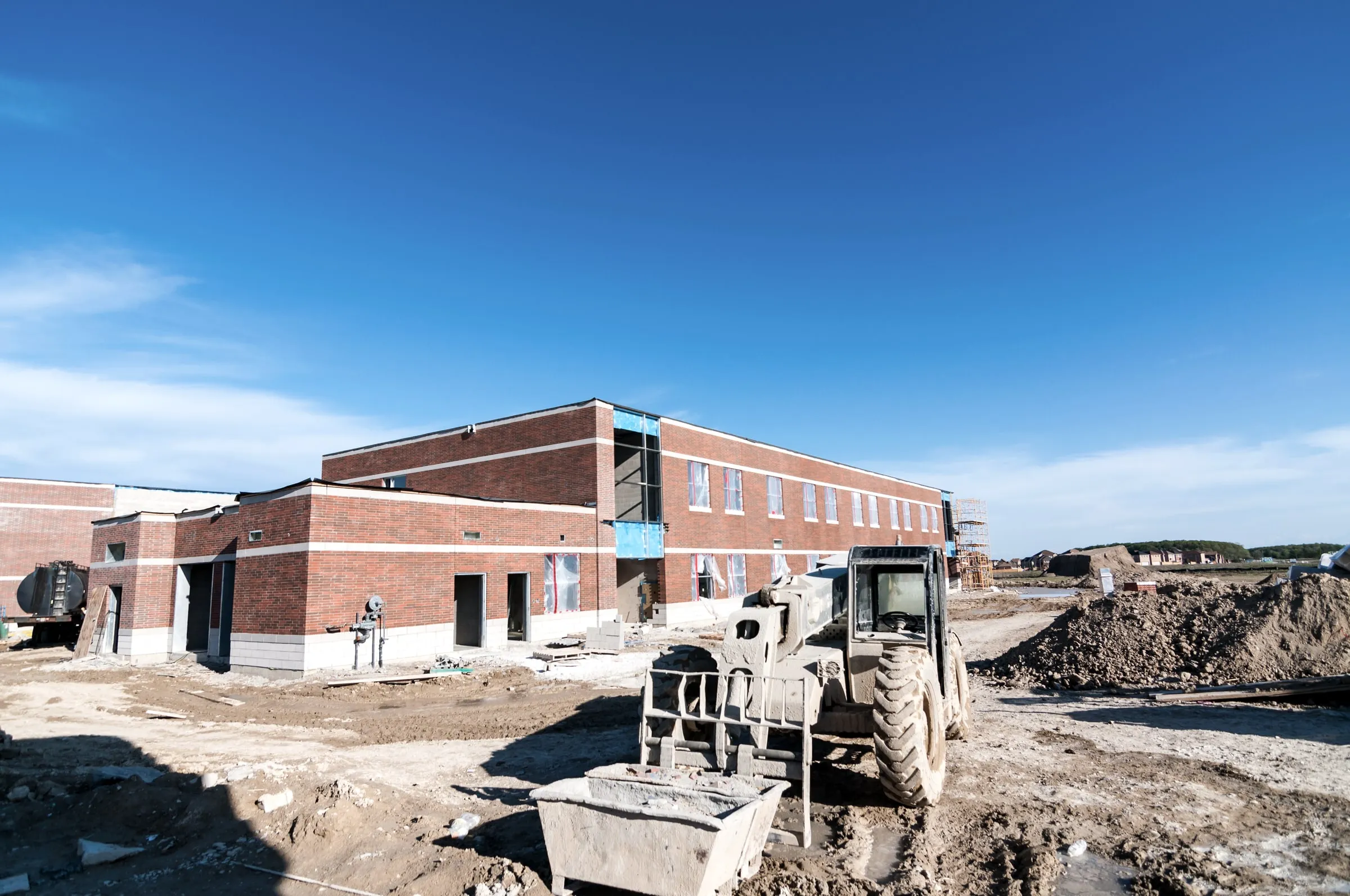 Two-story brick institutional building under construction with a telehandler forklift and building materials staged in front, under a clear blue sky