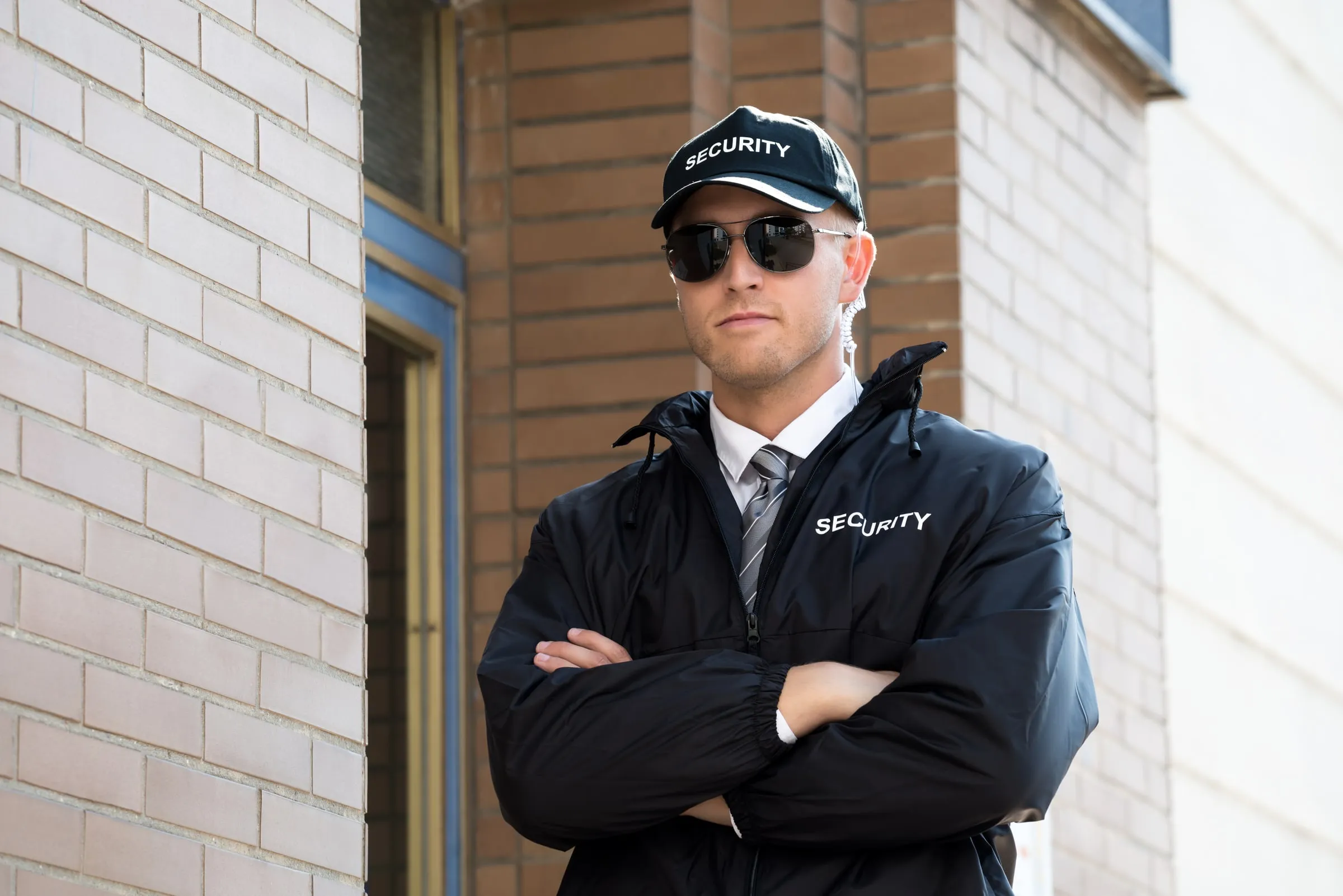 Security officer in a black SECURITY cap, sunglasses and a black SECURITY jacket standing with arms crossed beside a brick building entrance