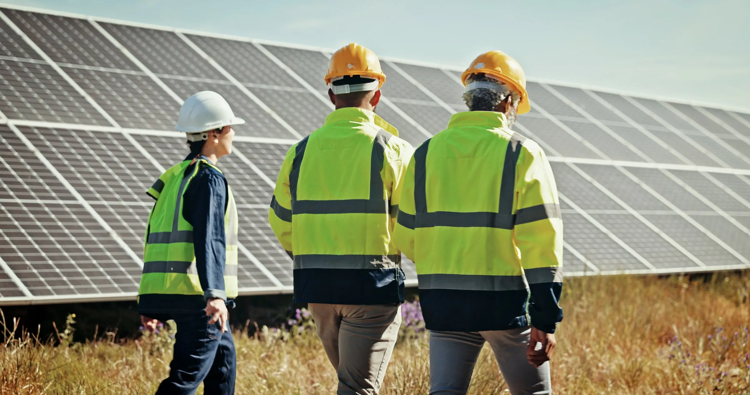 Three workers in hard hats and high-visibility vests walking through dry grass beside a row of ground-mounted solar panels on a sunny day