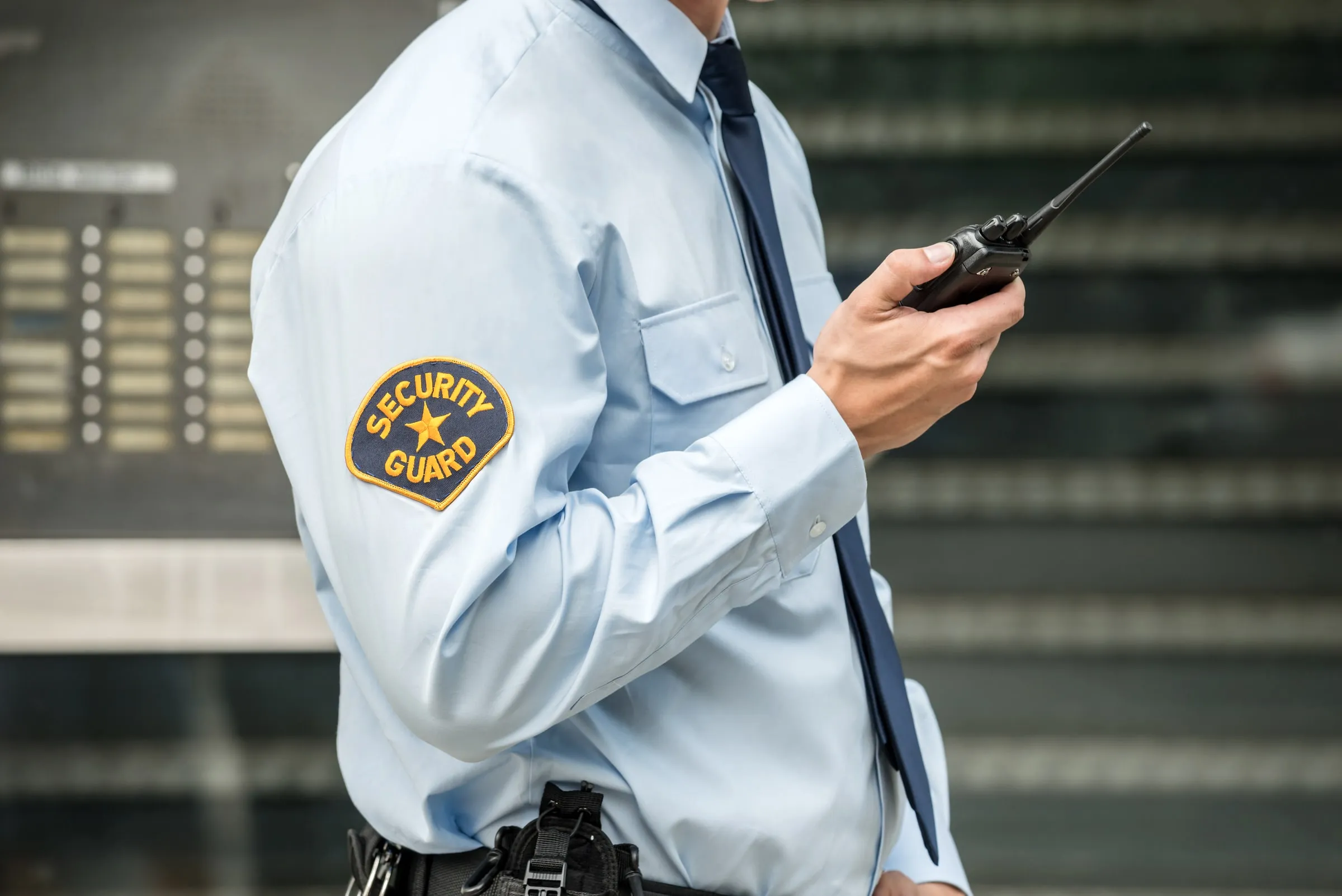 Close view of a security officer's shirt sleeve with a Security Guard patch, holding a handheld radio, standing in front of a glass building facade