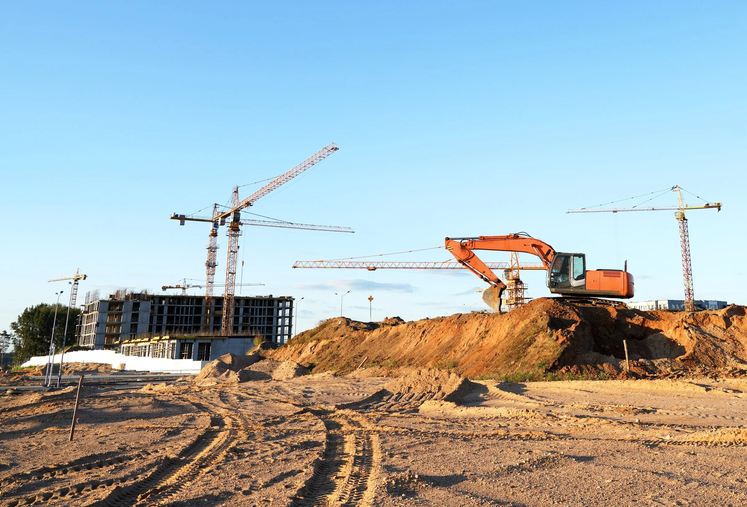 Orange excavator parked on a raised dirt mound at an empty construction site with no workers present, tower cranes and a mid-rise building under construction in the background