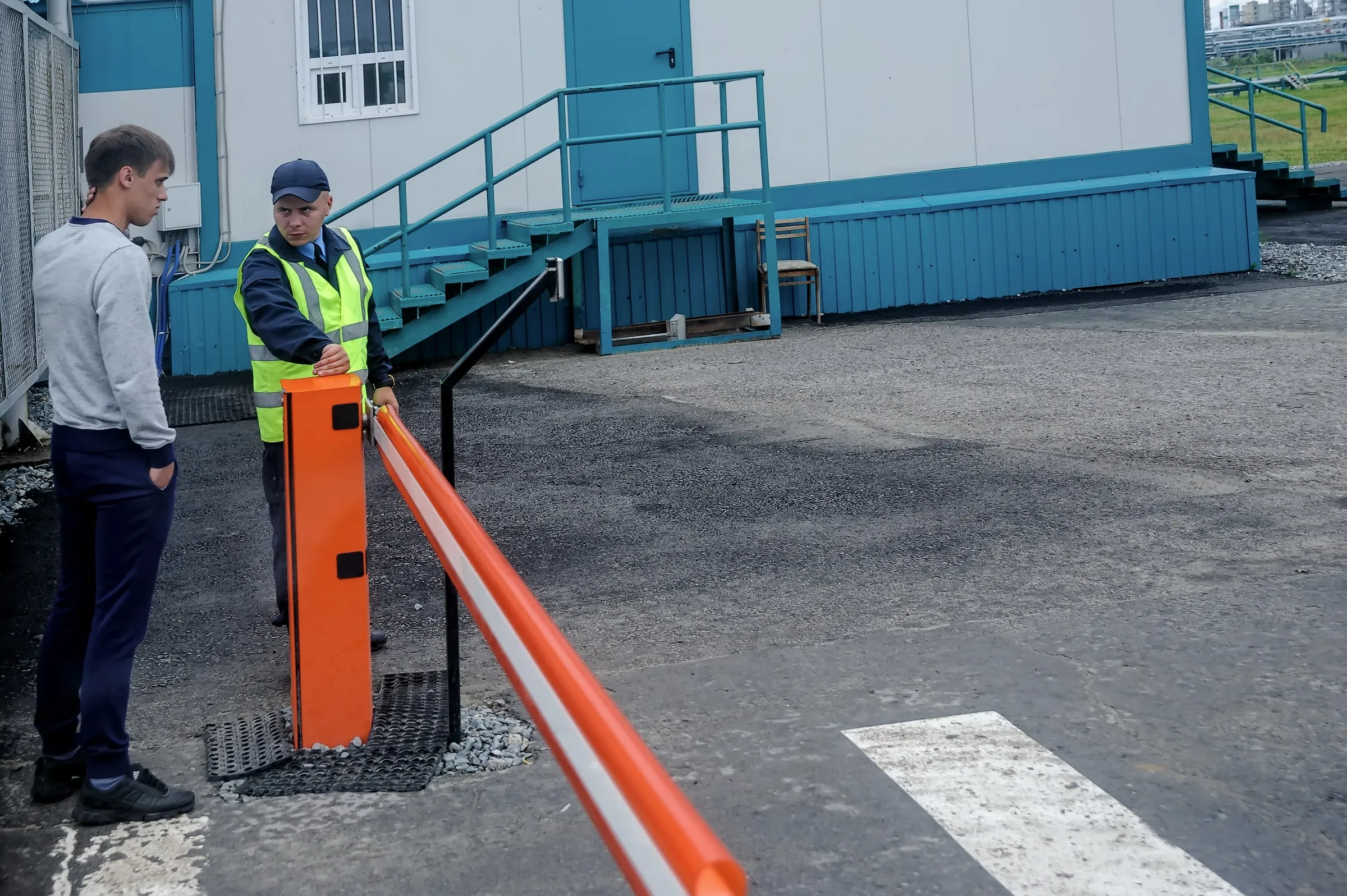 Security officer in a high-visibility vest and cap raising a vehicle gate arm at a facility entrance while a man waits beside him