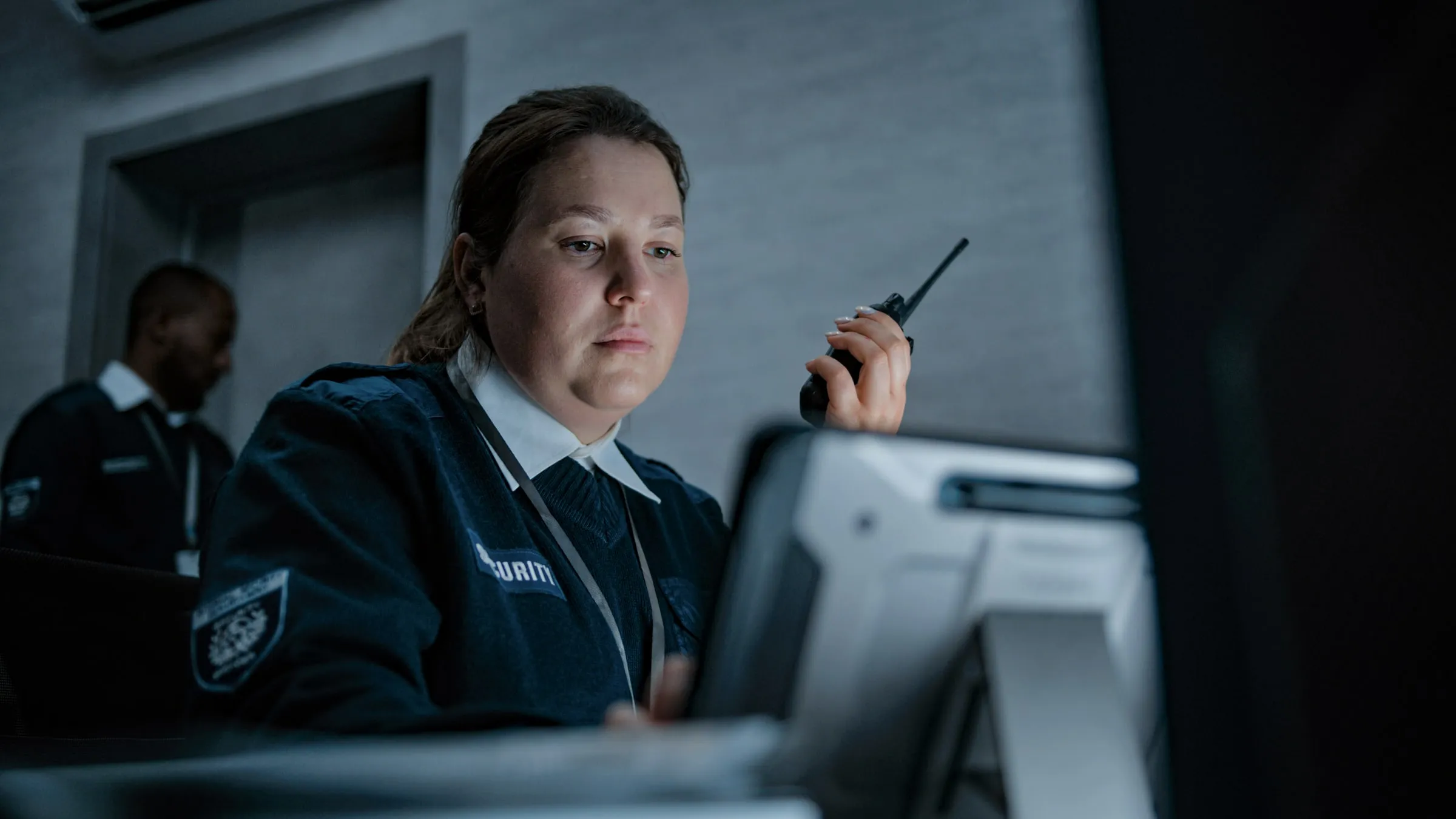 Security officer in uniform with a shoulder patch reading a monitor while holding a handheld radio to speak, another officer visible behind her